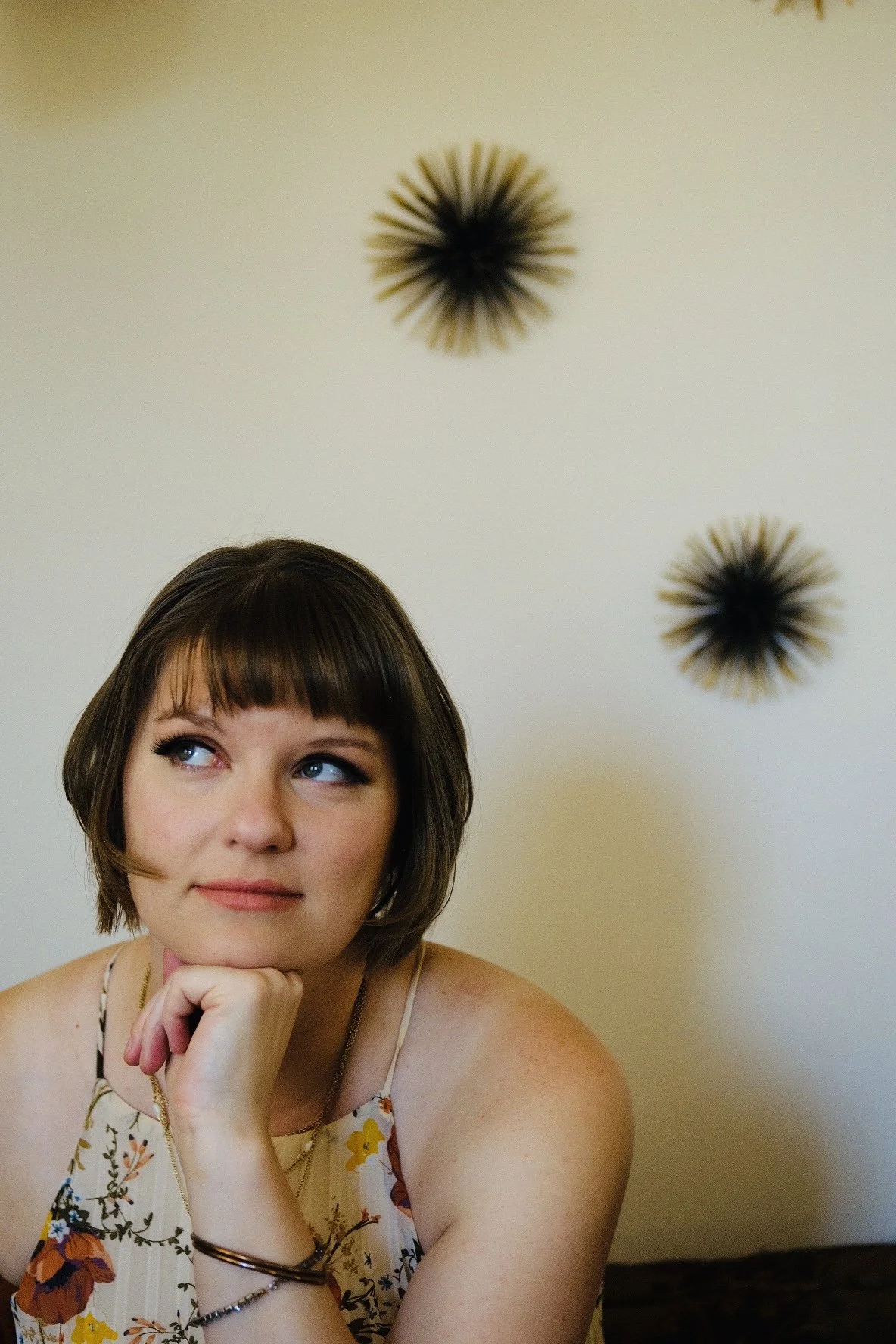 A woman with short brown hair and blue eyes resting her chin on her hand, wearing a floral top with thin straps, sitting in front of a plain white wall with two black sunburst wall decorations.