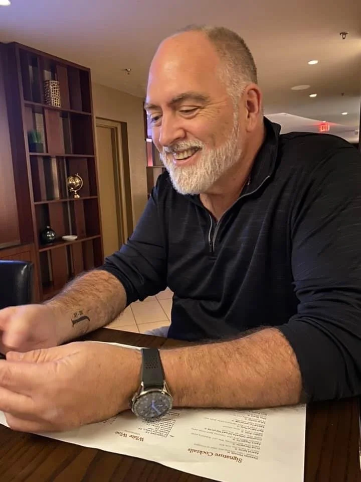 A middle-aged man with a beard and short hair, smiling, is seated at a table in a restaurant or cafe. He is wearing a black long-sleeve shirt, a watch, and has a tattoo on his forearm. He is looking at a smartphone in his hands, with a menu on the table in front of him.