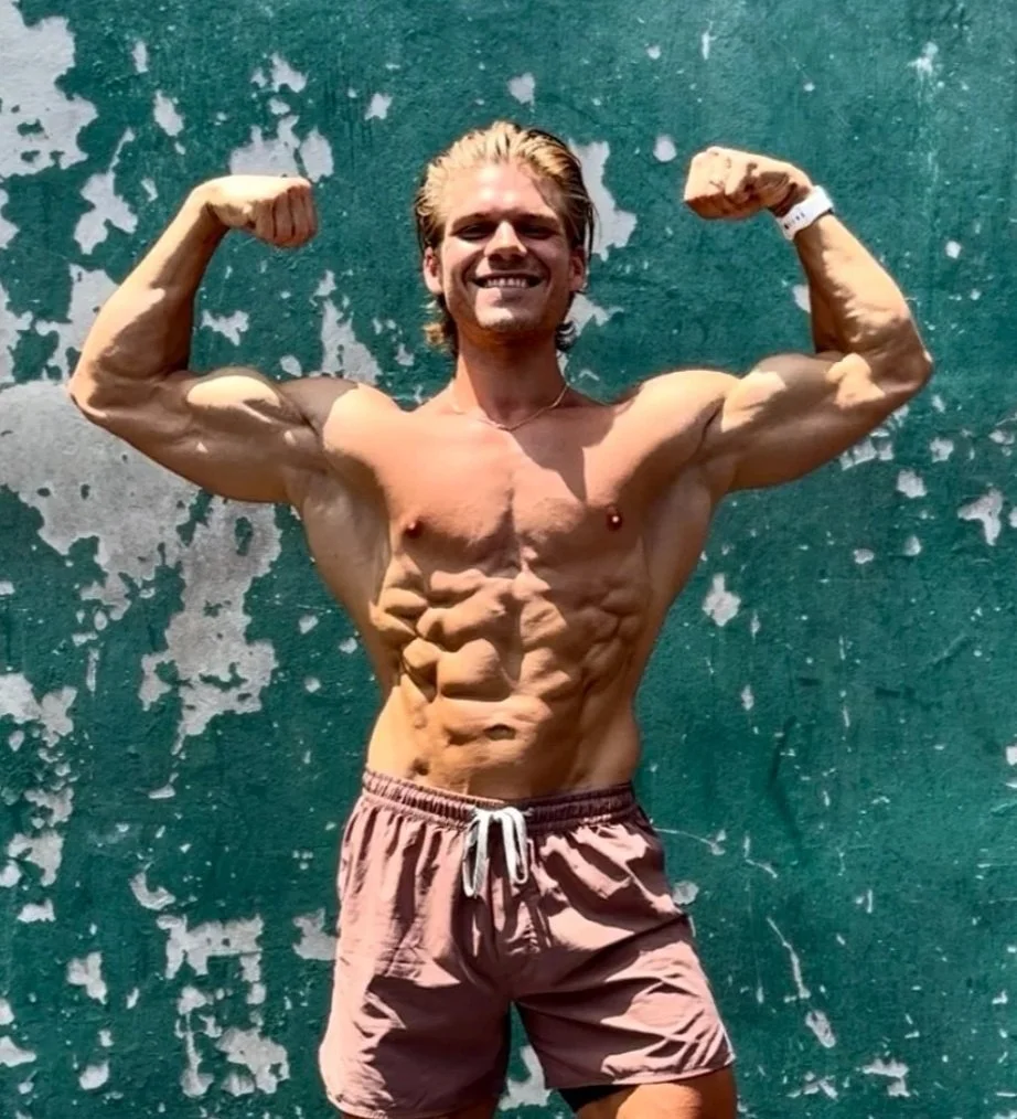 A shirtless man flexing his muscles and showing defined abs and biceps in front of a distressed green wall.