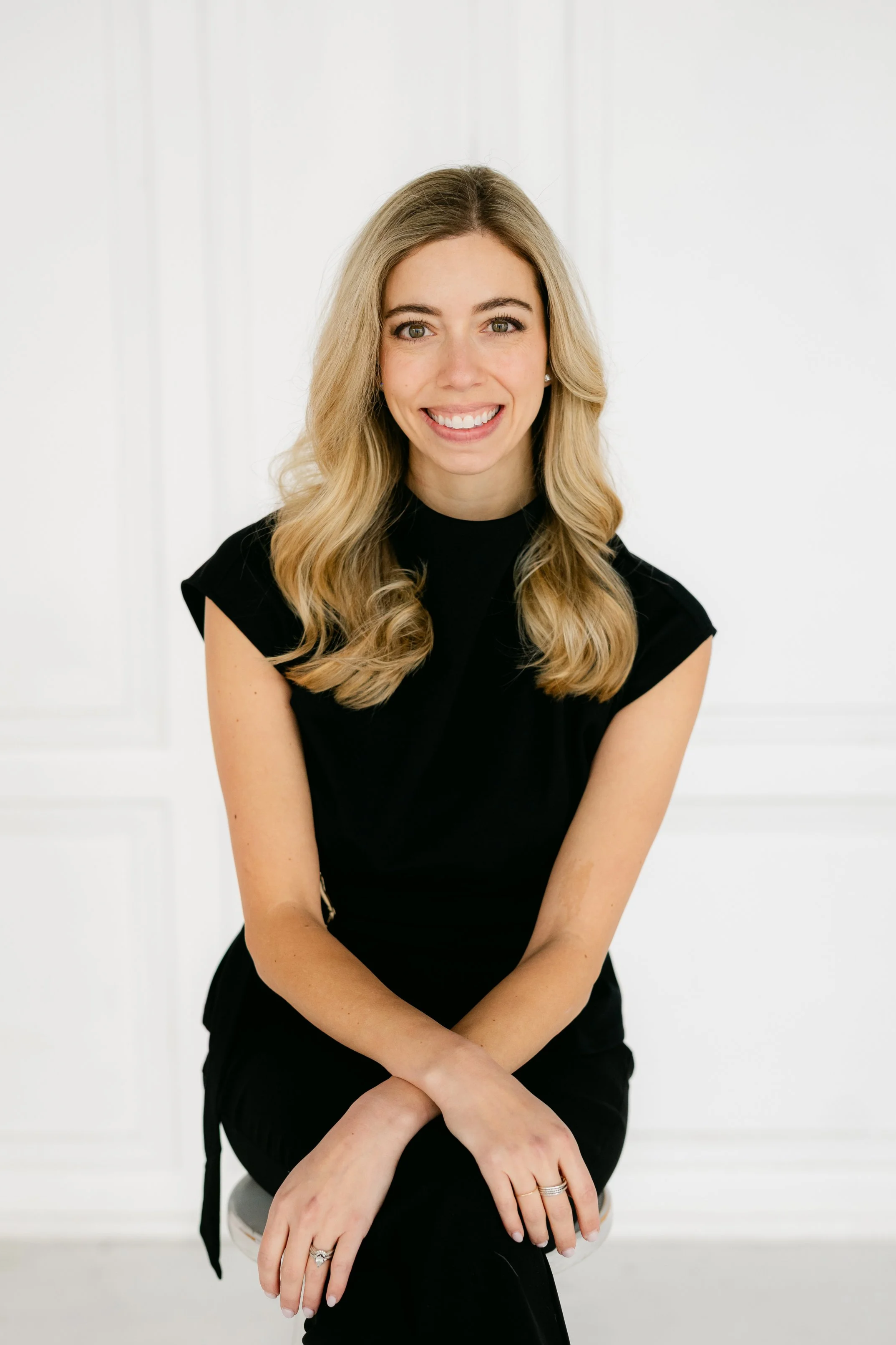 A woman with long blonde hair, wearing a black sleeveless top, sitting on a stool, smiling, in front of a white background.