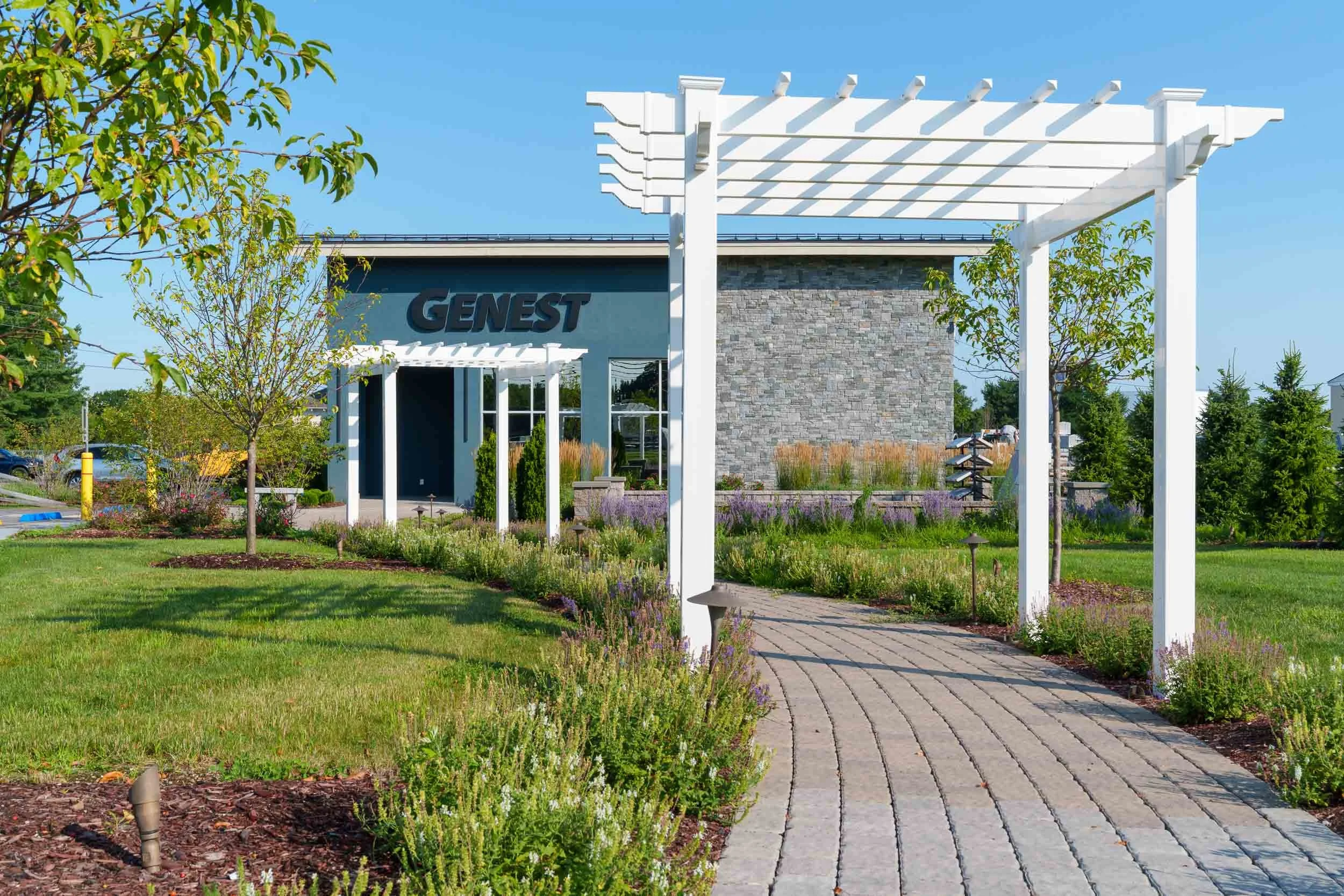 Exterior view of a modern building with a blue sign reading 'GENEST', surrounded by landscaped greenery including small trees, shrubs, and a paved walkway under a white pergola structure, all against a clear blue sky.