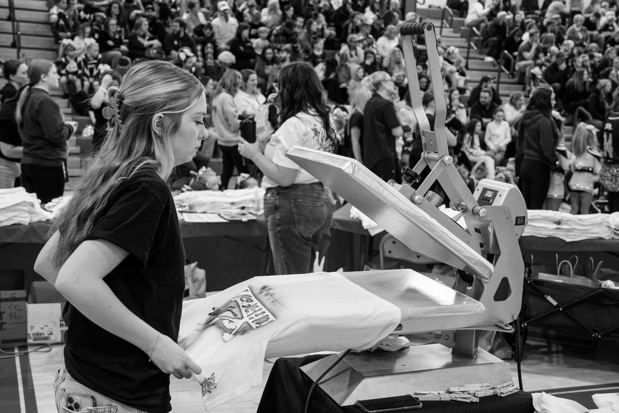 A young woman using a heat press to cure airbrush paint, while working at a custom airbrush painting booth during a cheering competition.