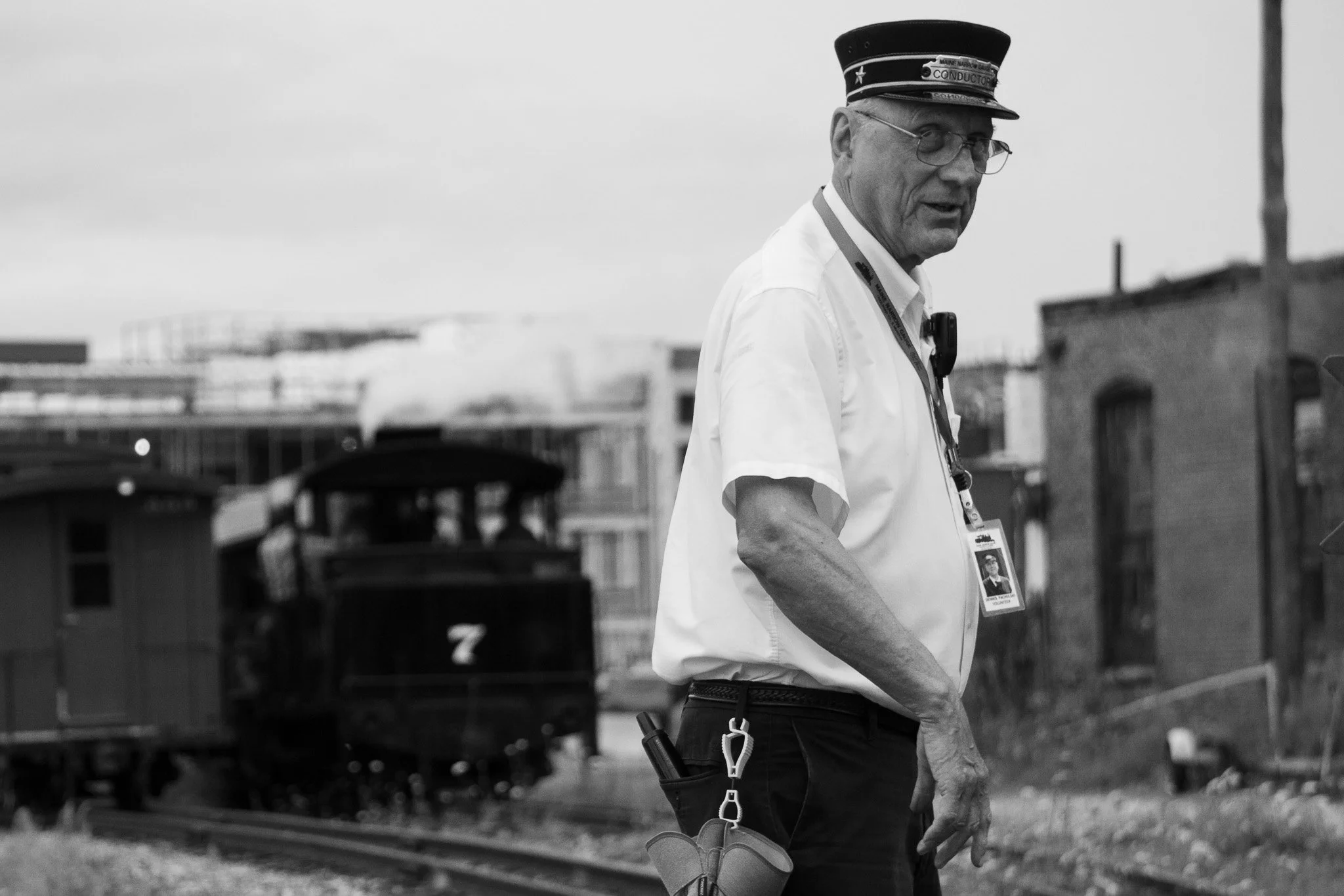 A man wearing a conductor's cap, glasses, and a white shirt hard hat stands outdoors near a train track. He has a badge around his neck and equipment attached to his belt. The background shows industrial buildings and a cloudy sky.