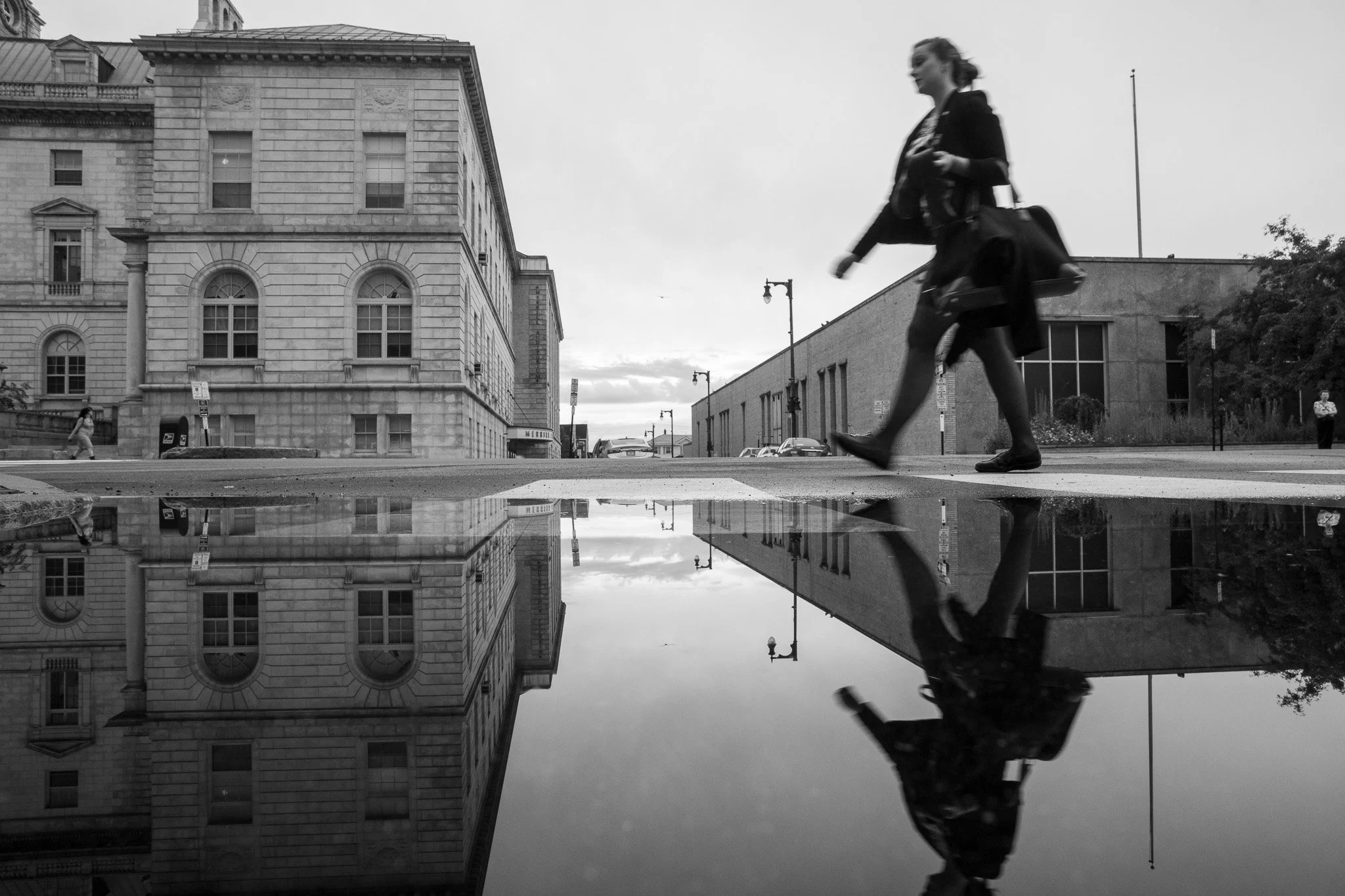 Female walking through frame, and reflected in a puddle after a summer rain near Portland City Hall, Maine.
