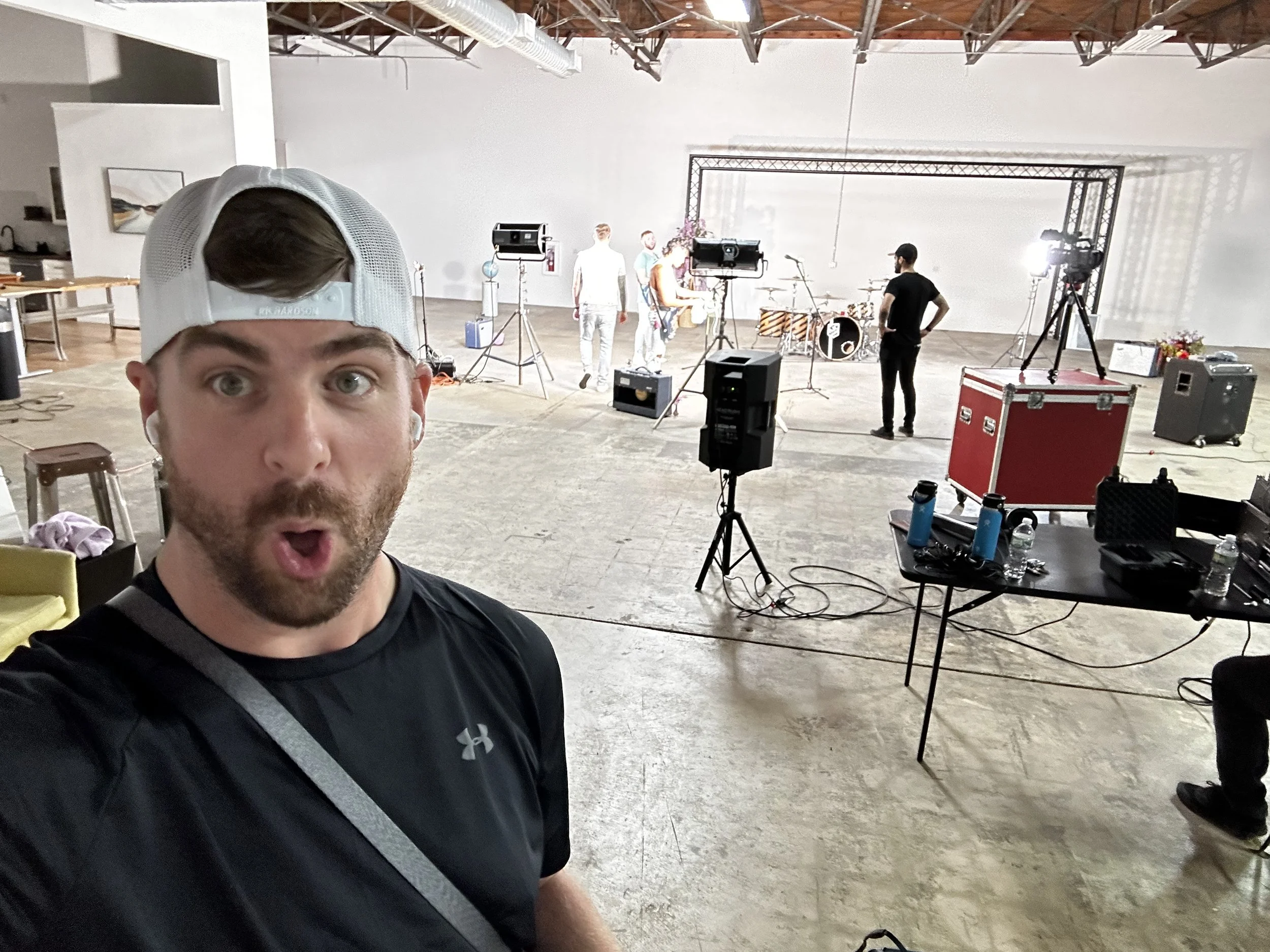 A man wearing a black Under Armour shirt and a white baseball cap is taking a selfie with his mouth open in surprise or excitement. Behind him, there is a stage setup with musical instruments and equipment in a large, spacious, industrial-style room with a high ceiling and exposed beams. There are several people on the stage, and professional lighting and camera equipment are visible.