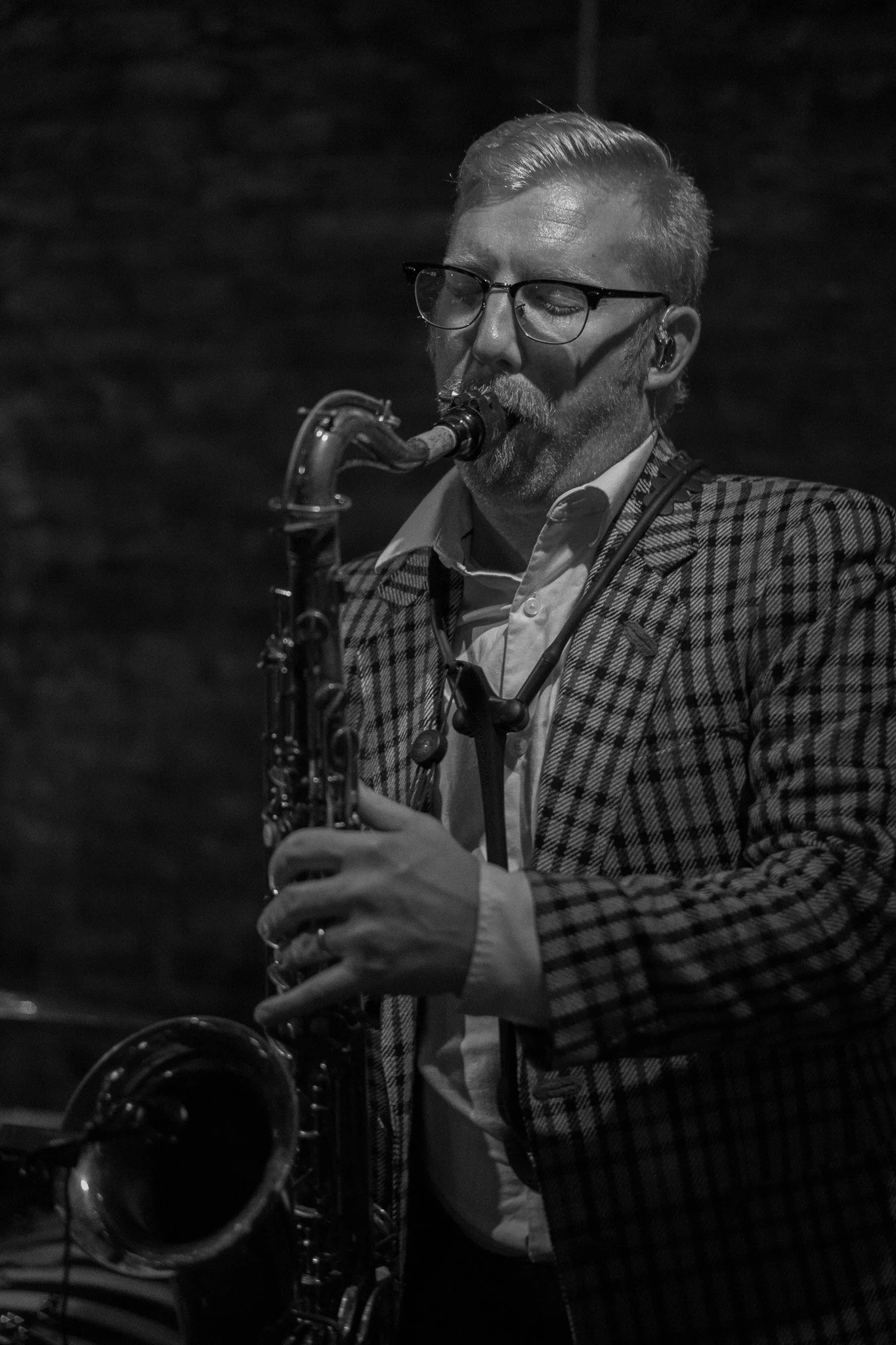A man wearing glasses and a checkered blazer, playing a saxophone in a dimly lit setting.