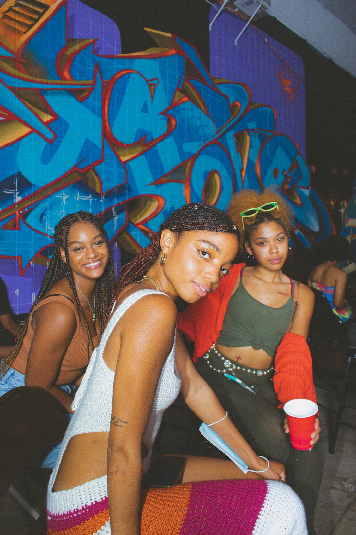 Photo of three Black women seated looking to camera in front of graffiti mural