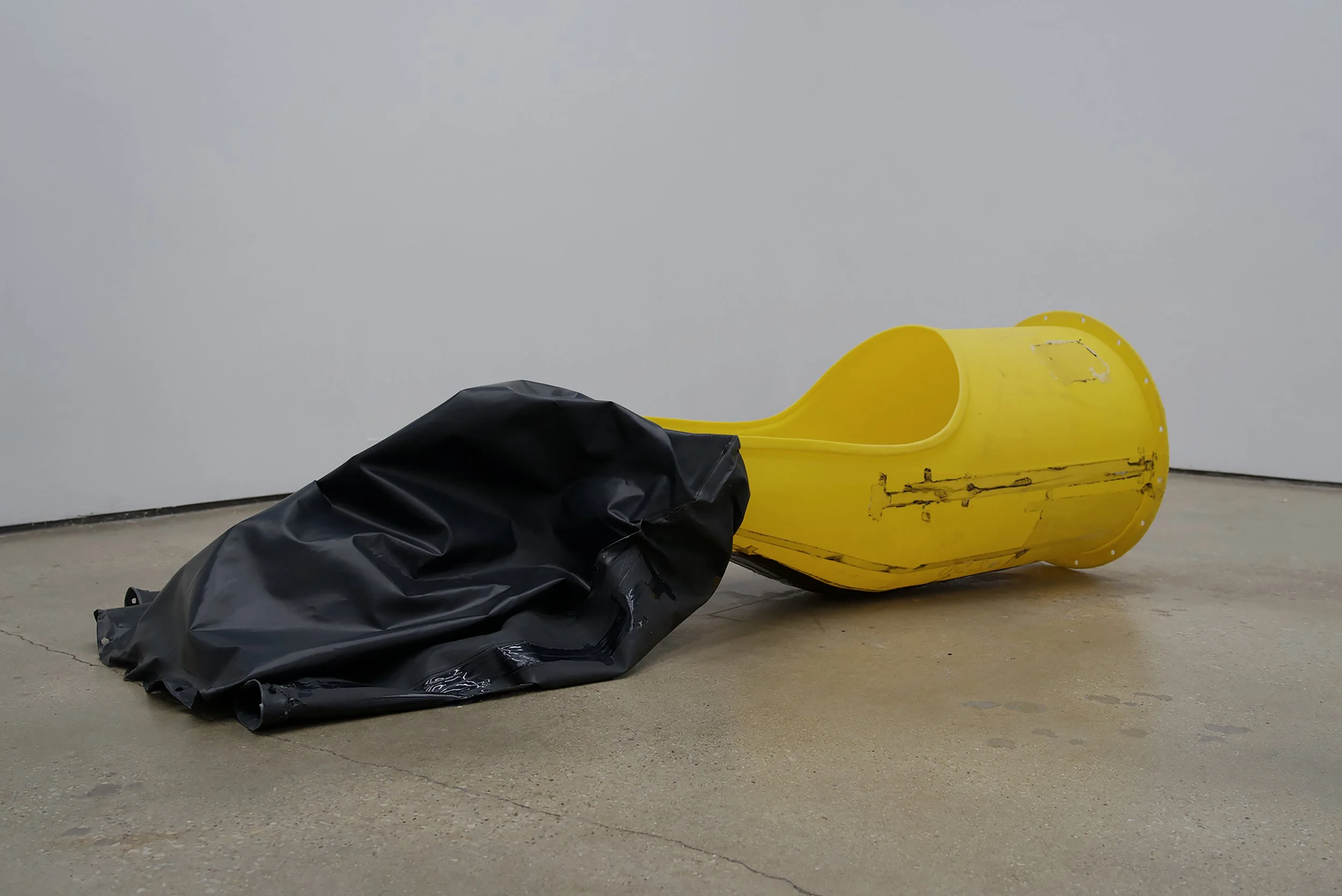 A yellow plastic or fiberglass kayak with a black cover or tarp partially draped over it, resting on a concrete floor against a white wall.