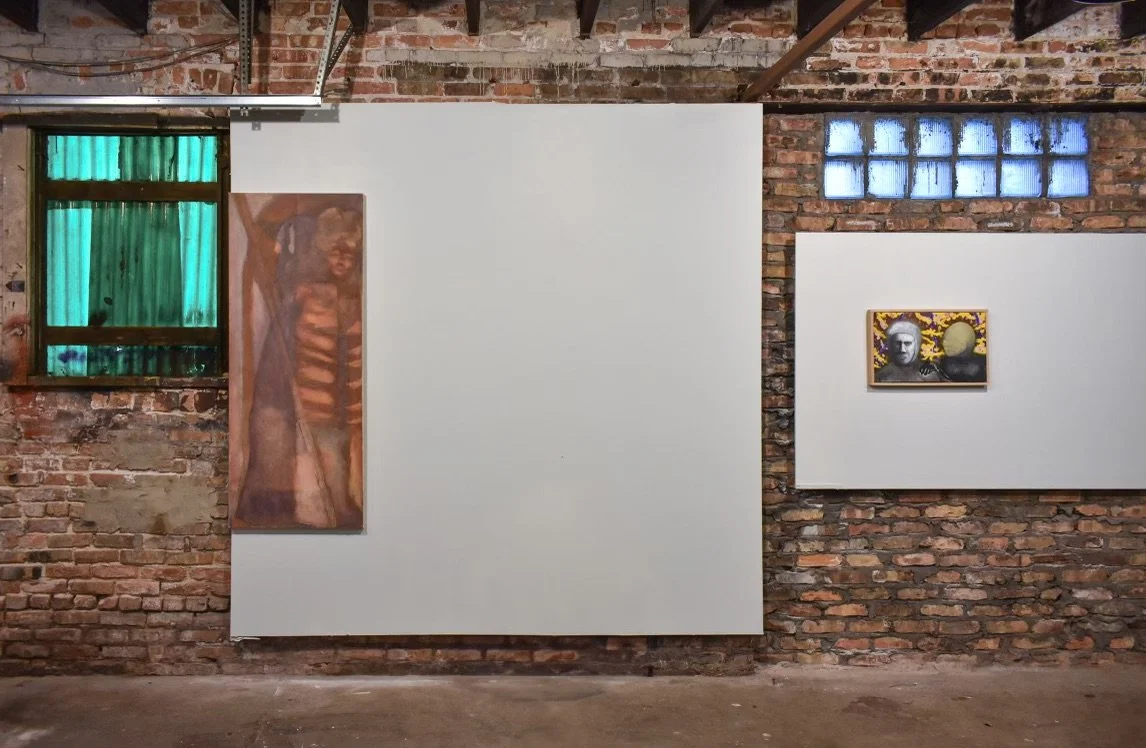 Exhibition gallery with brick walls, featuring two artworks; one blurry painting of a person with a striped shirt on a white panel, and a black-and-white portrait of a woman with a yellow and purple background.