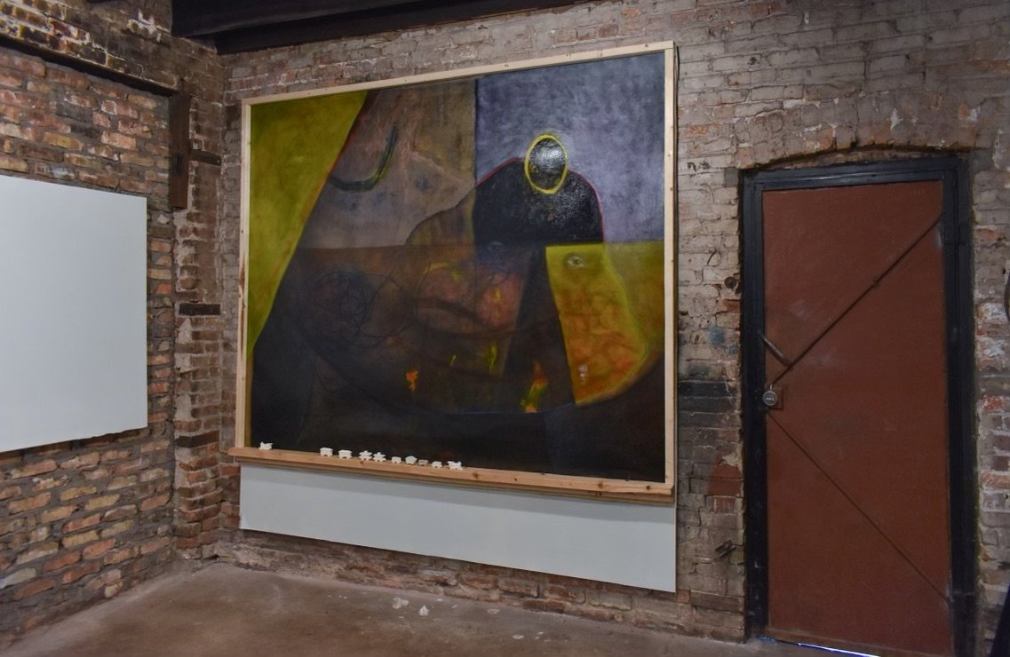 Abstract painting on brick wall with dark, muted colors, a large black circle, and yellow accents.