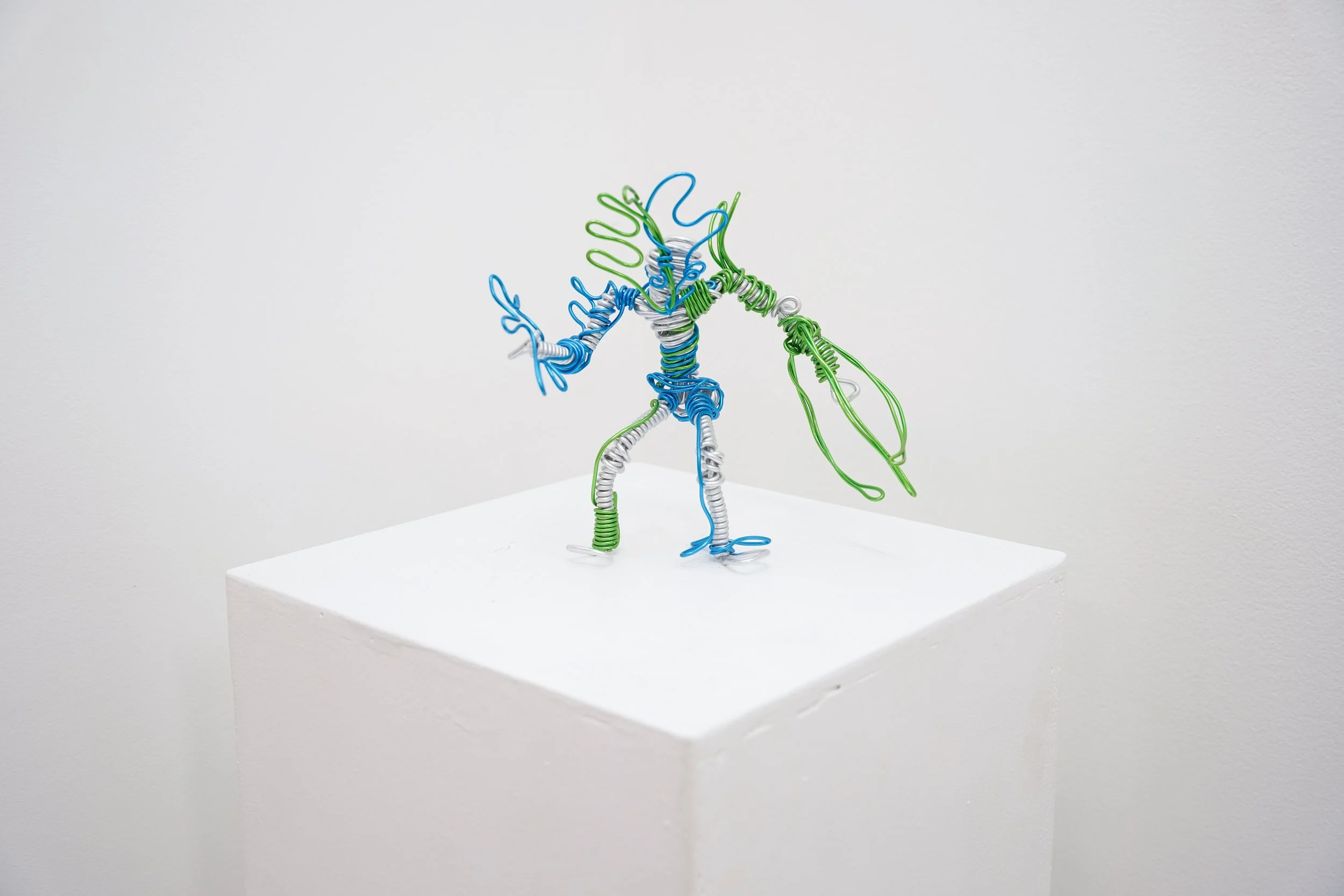 Colorful wire sculpture of a humanoid figure with gloves, standing on a white pedestal against a plain wall background.