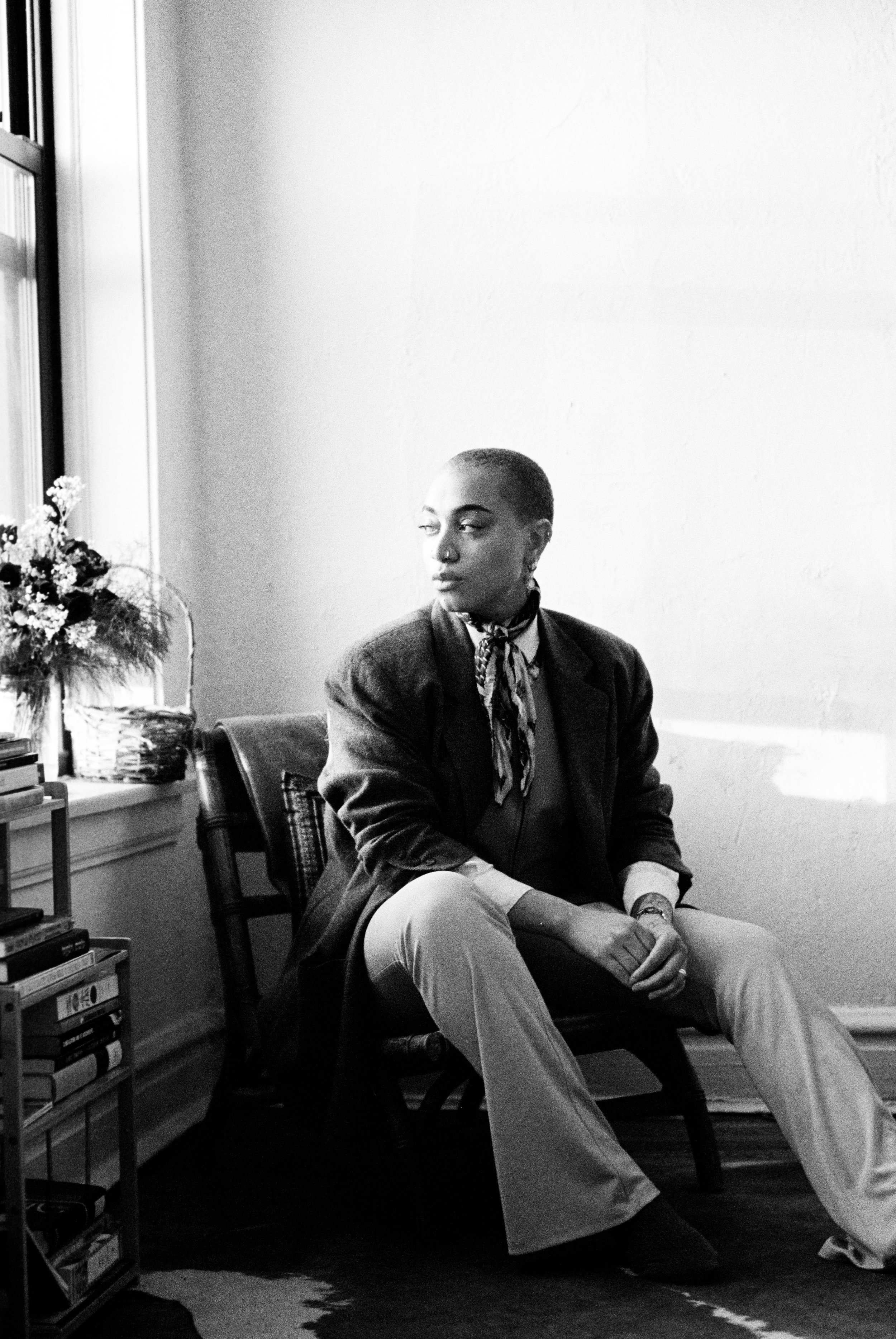 Black and white photo of a woman with short hair, sitting on a chair by a window, looking to the side. She is wearing a blazer, a patterned scarf, and light-colored pants. There are books and flowers on a nearby shelf.