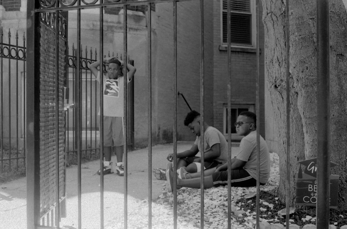 Photo by Isaiah Ward, 3 young Black boys two sitting, one with glasses the other with his eyes to the ground, next to a third boy standing while looking off into the distance. All three captured on 35 mm film from behind a fence.