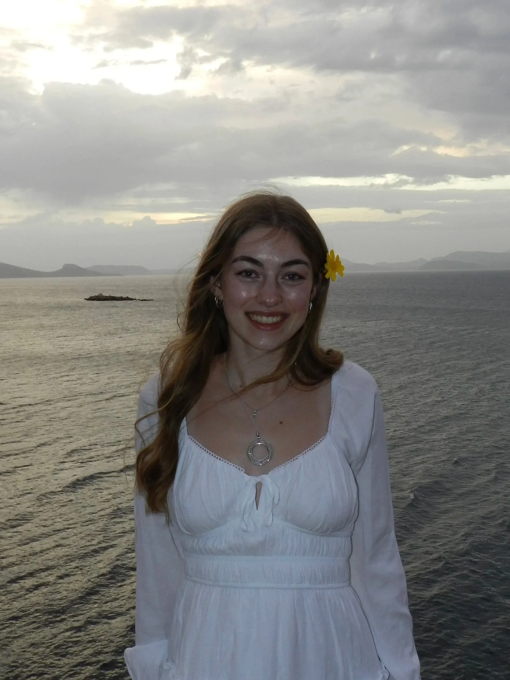 A young woman with long, wavy red hair smiling at the camera, standing outdoors near the ocean with hills and clouded sky in the background. She is wearing a white dress, silver jewelry, earrings, and a yellow flower in her hair.