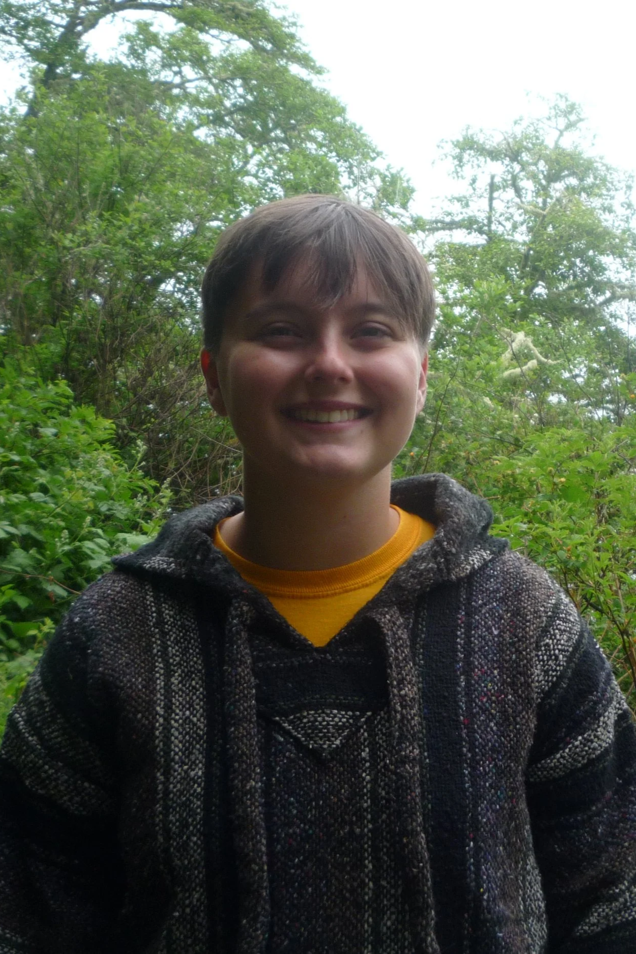 Smiling young person with short hair, wearing a patterned hoodie and yellow shirt, standing outdoors among green trees and bushes.