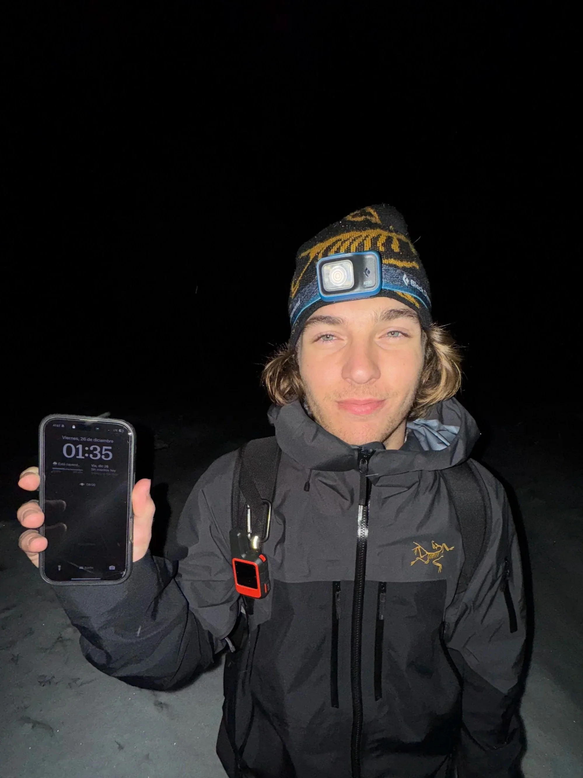 A man wears clear round glasses, has dark curly hair, a beard, and a nose ring. He is dressed in a dark sweater and is seated indoors with blurred background decor.