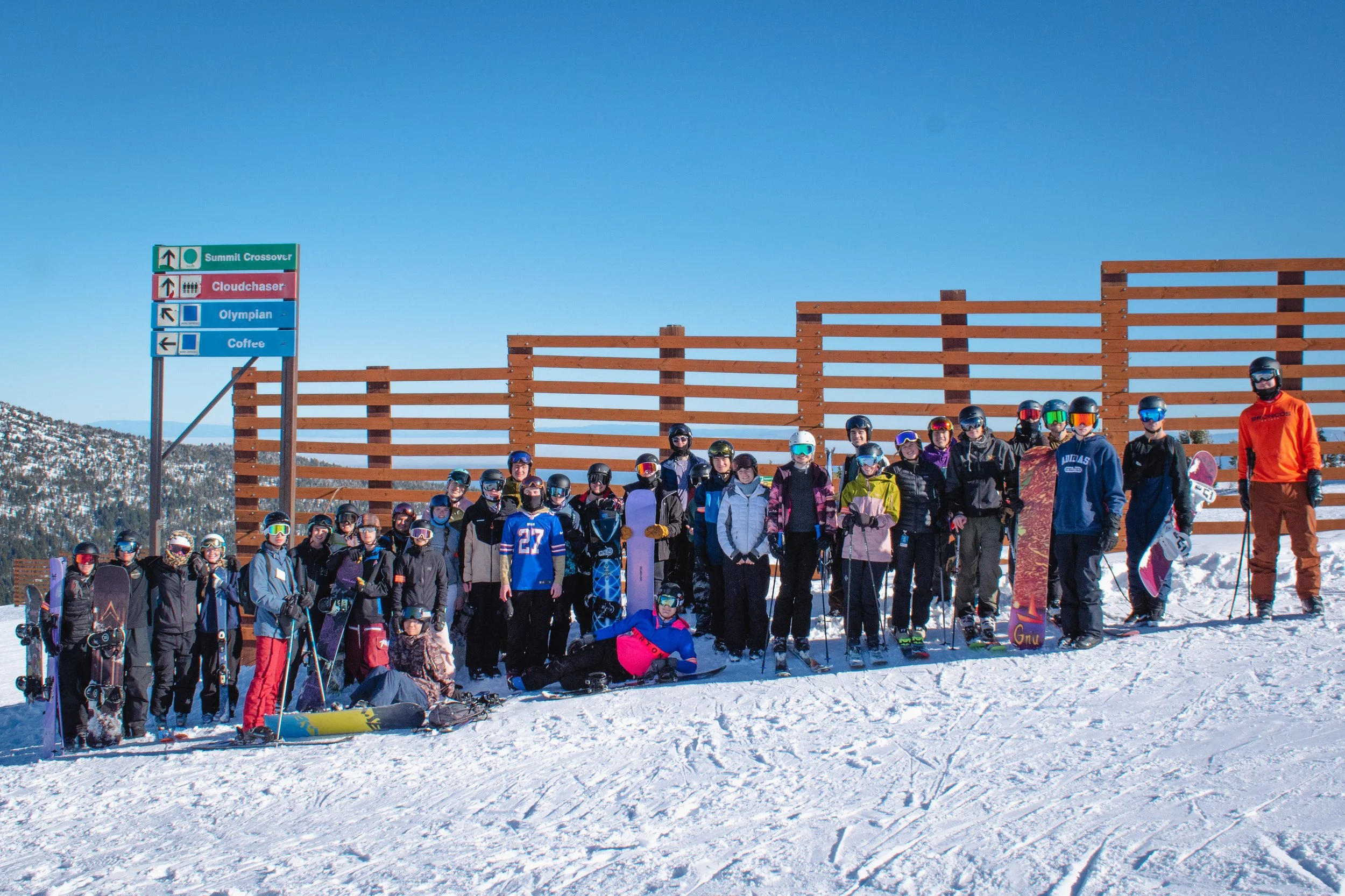 Group of skiers and snowboarders posing on a snowy mountain with clear blue sky, wearing helmets and goggles, with ski equipment, in front of a wooden fence and a signpost with ski trail names.