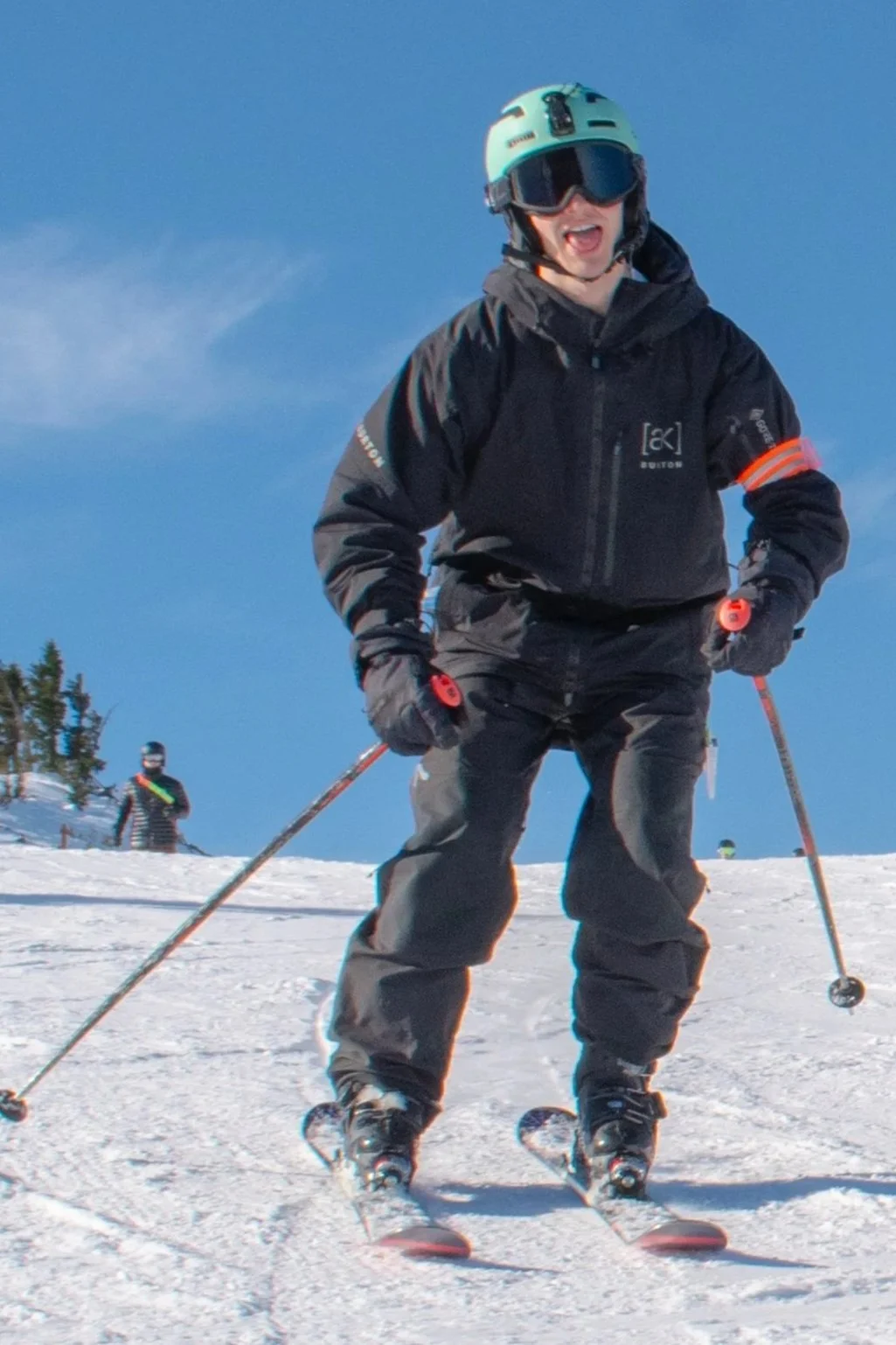 A young man wearing ski goggles and a black winter jacket, sitting inside a ski lift with a snowy mountain and ski resort in the background.