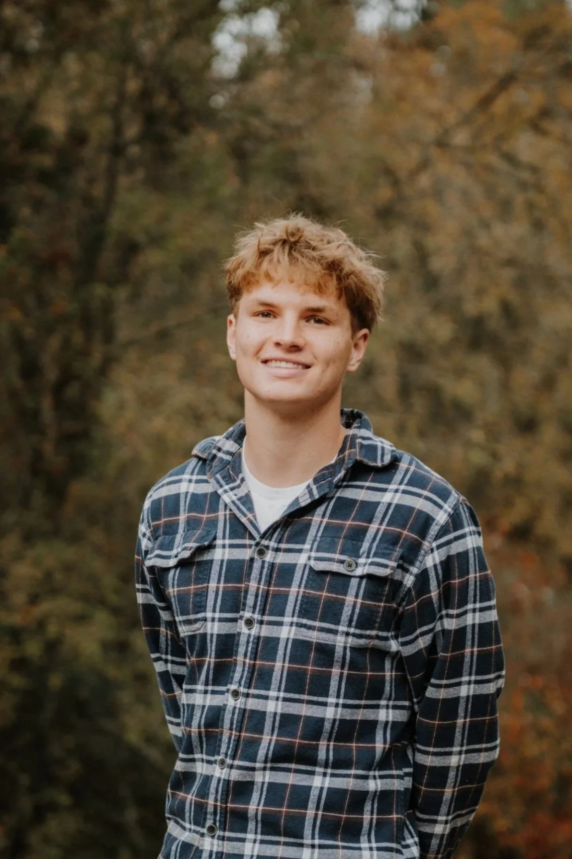 Young man with curly light brown hair smiling while standing outdoors in a natural setting with trees and autumn foliage.