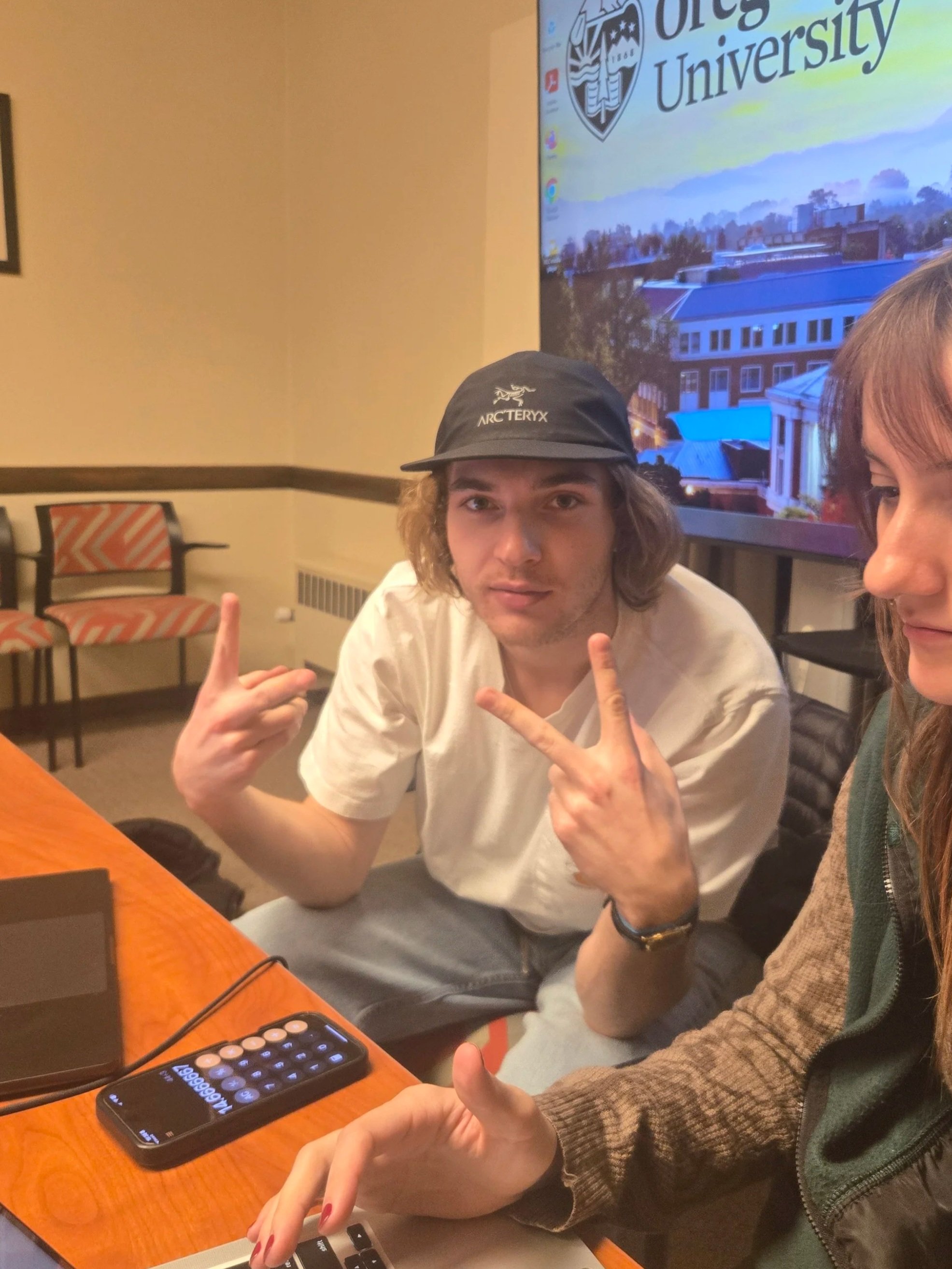 A man wears clear round glasses, has dark curly hair, a beard, and a nose ring. He is dressed in a dark sweater and is seated indoors with blurred background decor.