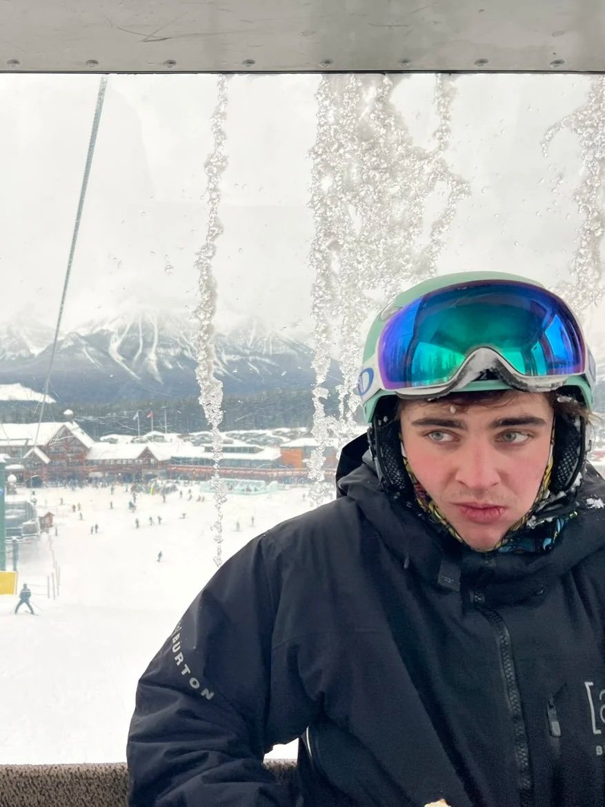A young man wearing ski goggles and a black winter jacket, sitting inside a ski lift with a snowy mountain and ski resort in the background.