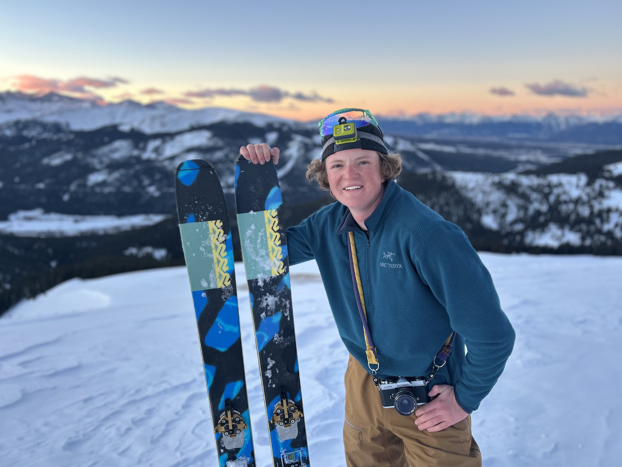 Young man standing on snow-covered mountain at sunset, smiling, holding skis, wearing a blue Arc'teryx jacket, a helmet with a headlamp, and a camera around his neck, with snow-capped mountains and a valley in the background.