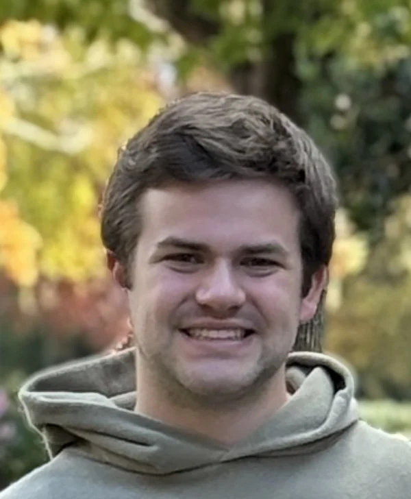 A young man smiling outdoors with trees in the background.