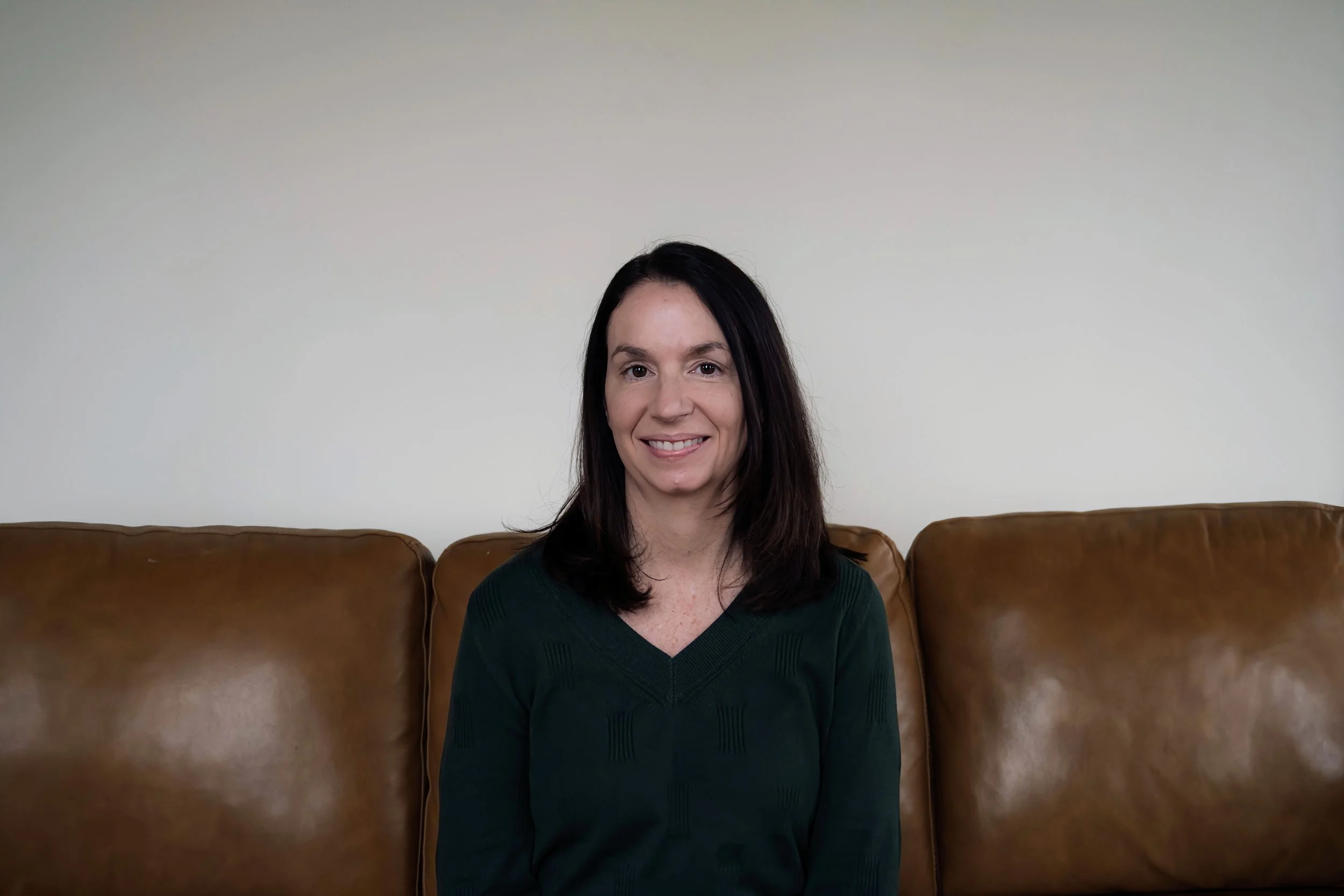 A woman with dark brown hair and a green sweater sitting on a brown leather couch in front of a plain white wall, smiling at the camera.