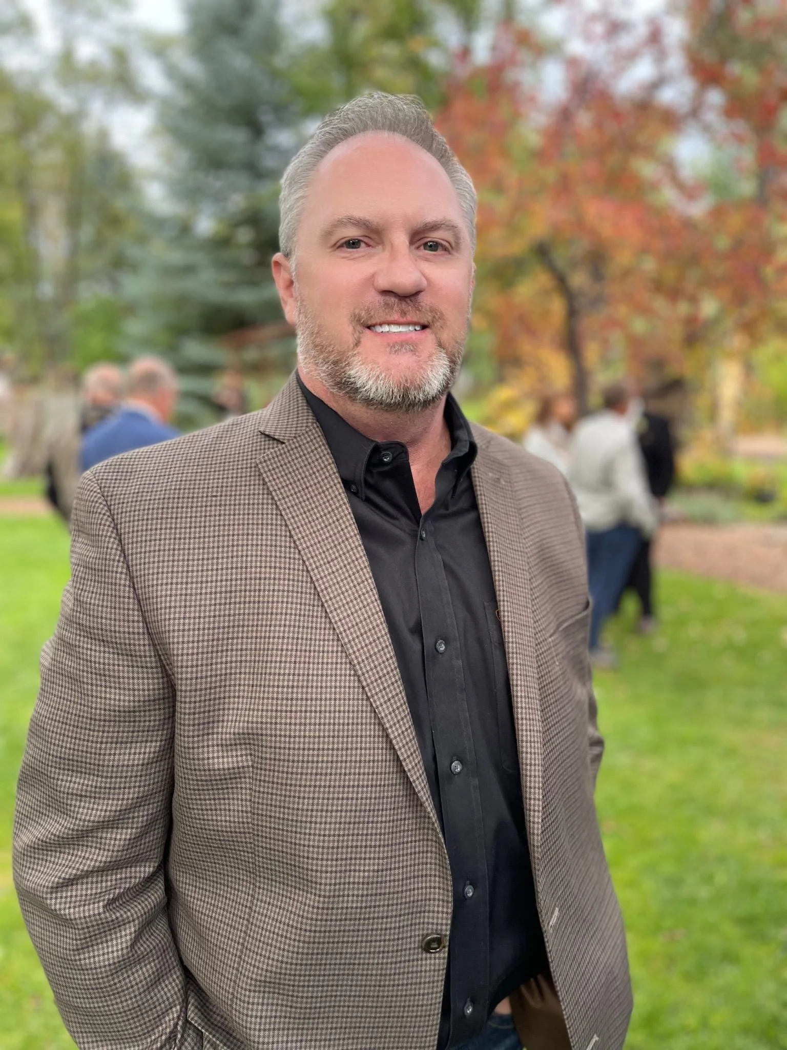 Man in a brown checkered blazer smiling outdoors with autumn trees and people in the background.