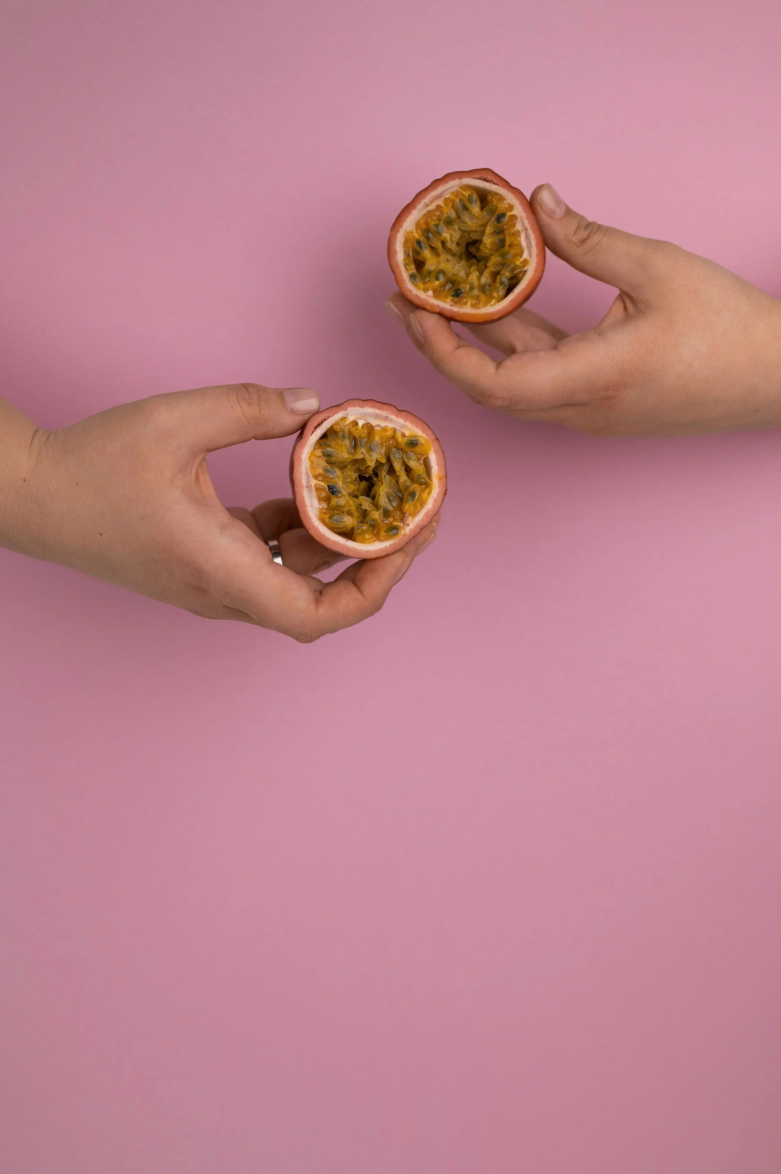 Two hands holding halved passion fruits with pink background.