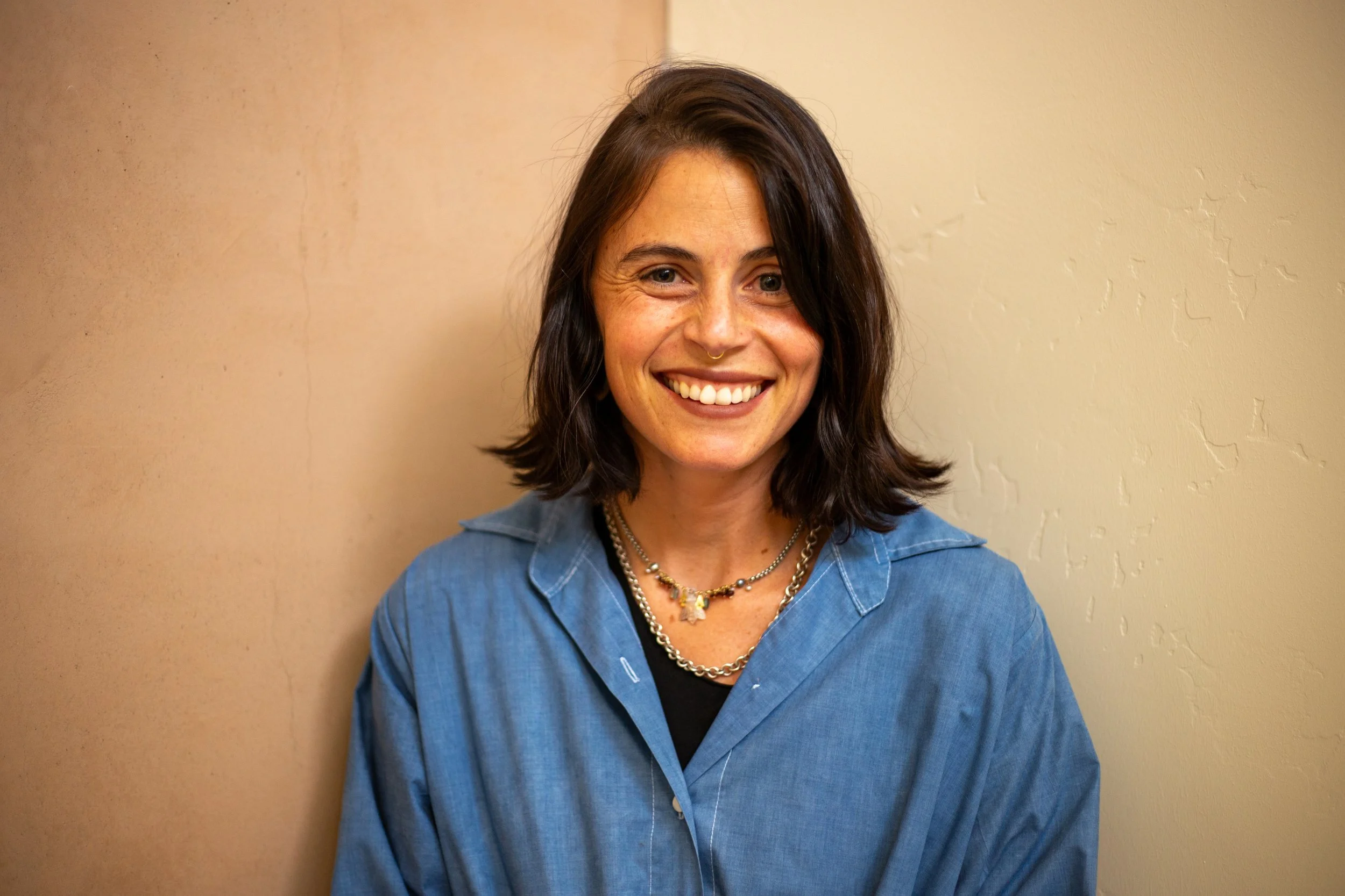 A woman with shoulder-length dark hair, wearing a blue jacket over a black top, smiling and standing in front of a beige textured wall.
