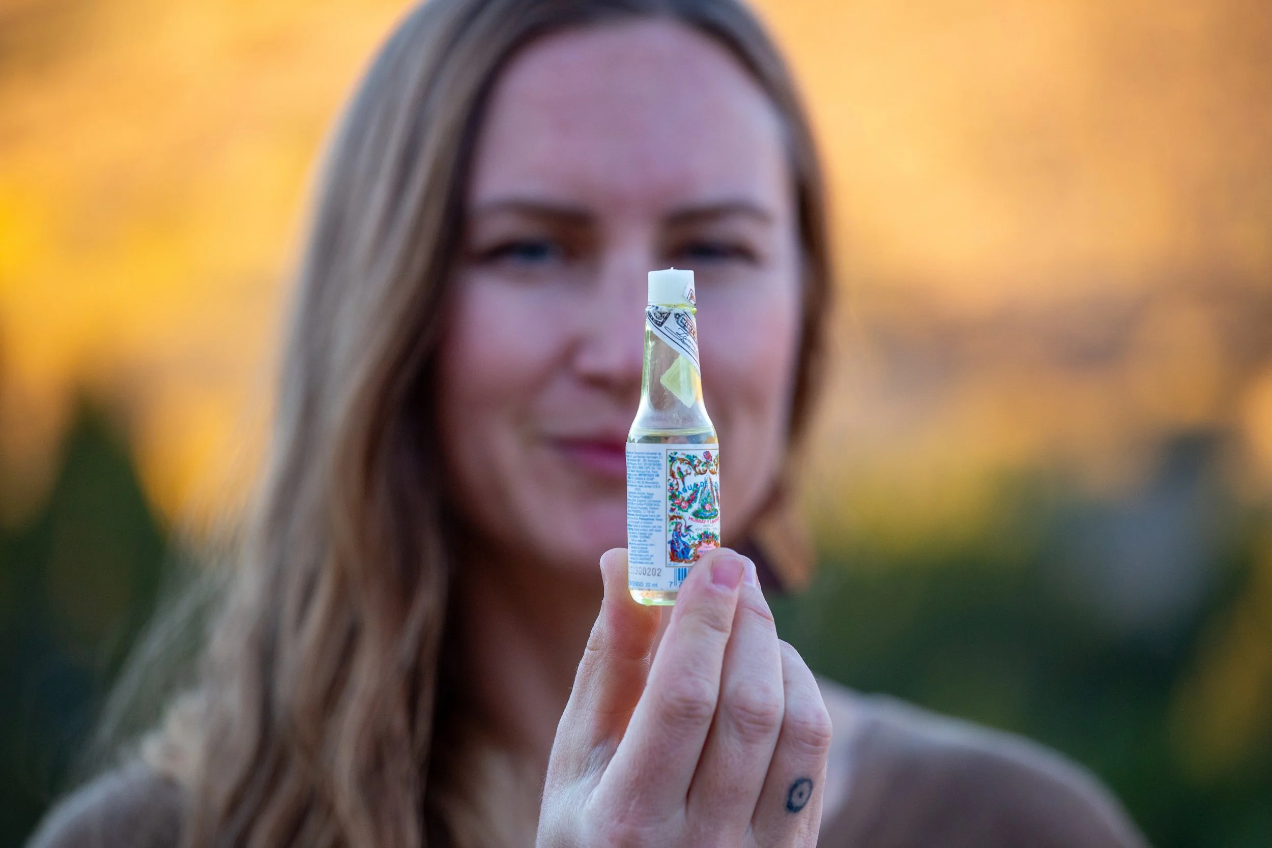 A woman with long red hair holding a small bottle of liquid, with a colorful label, in front of her face outdoors during sunset.