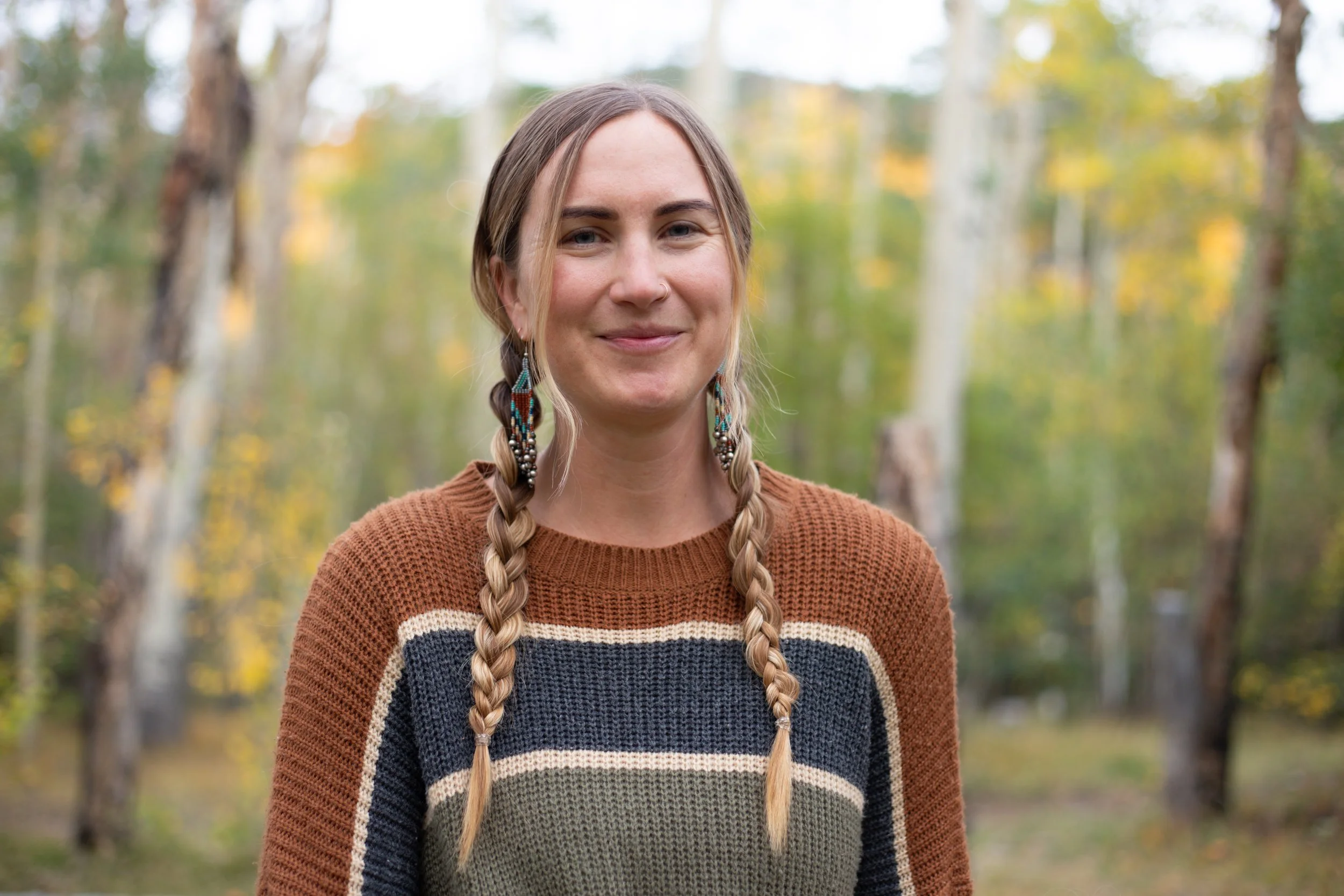 A woman with long braided hair, wearing a multicolored striped sweater, standing outdoors with a blurred background of trees with fall foliage.