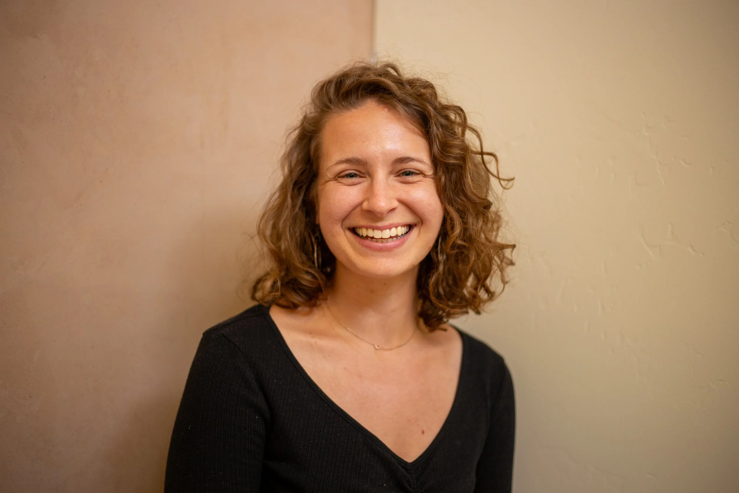 A woman with curly hair smiling, wearing a black top and a delicate necklace, standing against a beige wall.