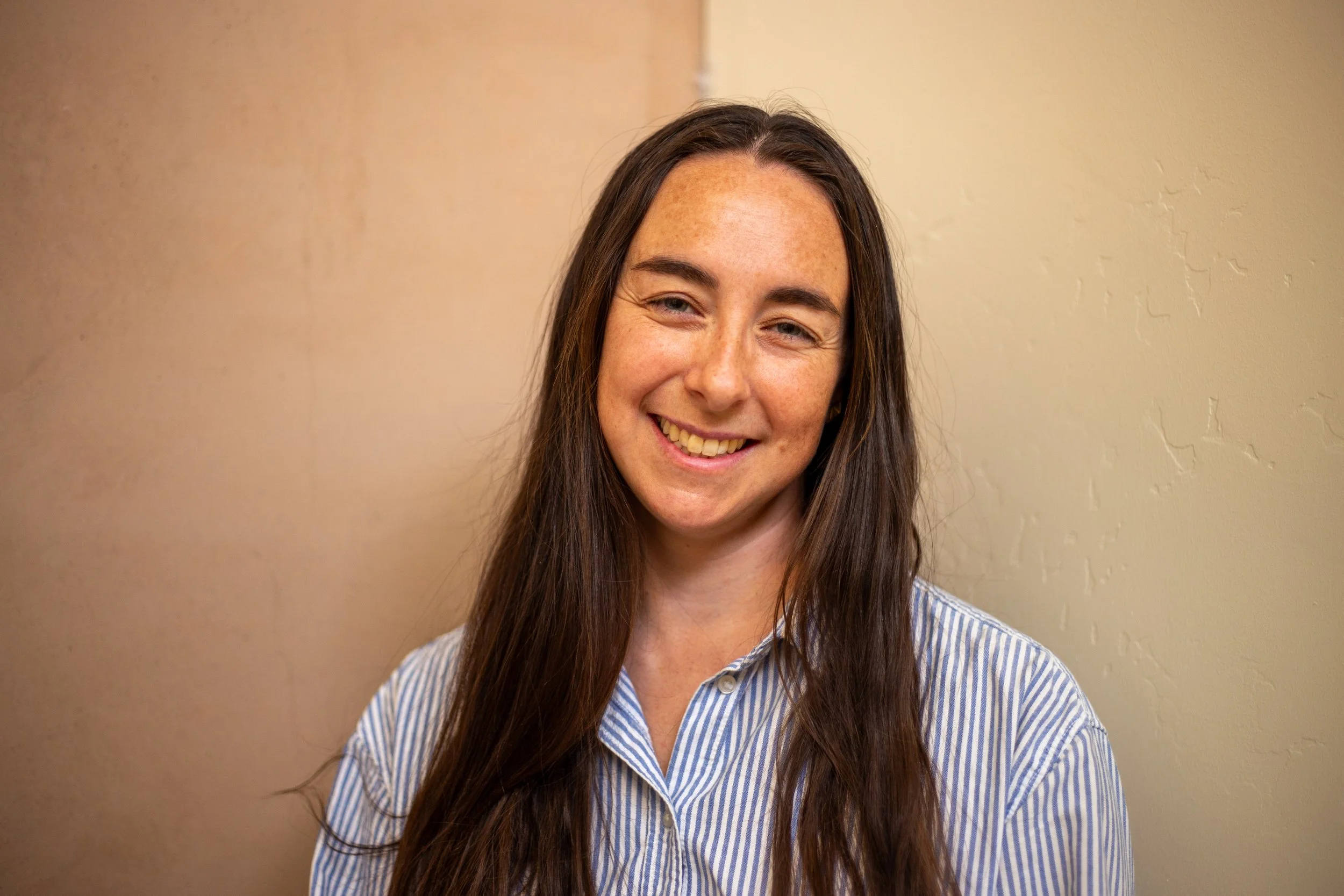 A young woman with long brown hair, freckles, and blue eyes smiling gently while wearing a blue and white striped shirt, standing against a beige wall.
