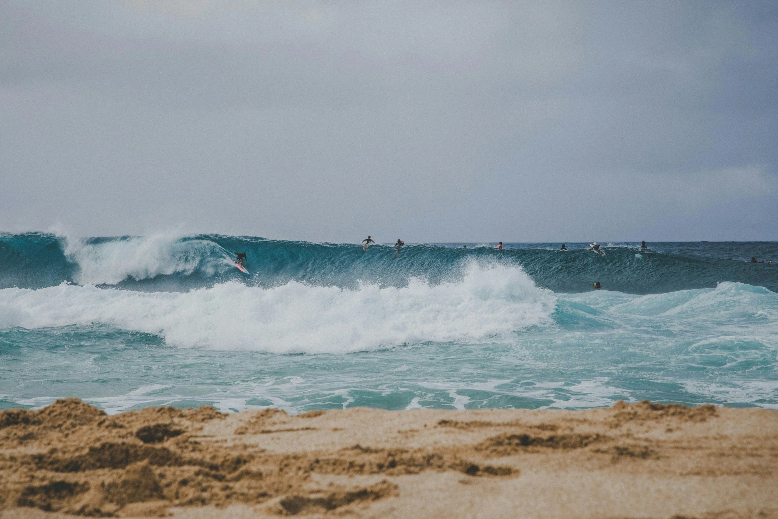 A beach scene with a surfer riding a wave, multiple surfers in the water, sandy beach in the foreground, and a cloudy sky.