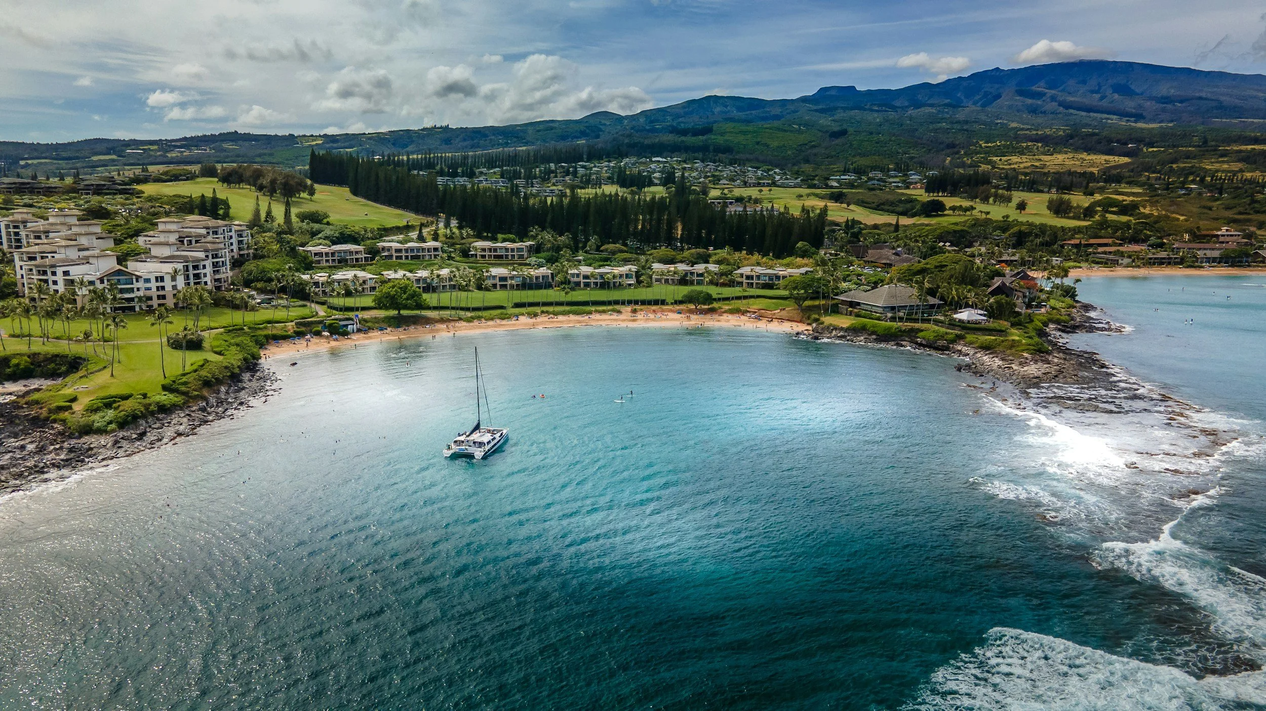 A coastal scene with a marina and luxury residences, a sandy beach with umbrellas, a sailboat anchored near the shoreline, and mountains in the background under partly cloudy skies.