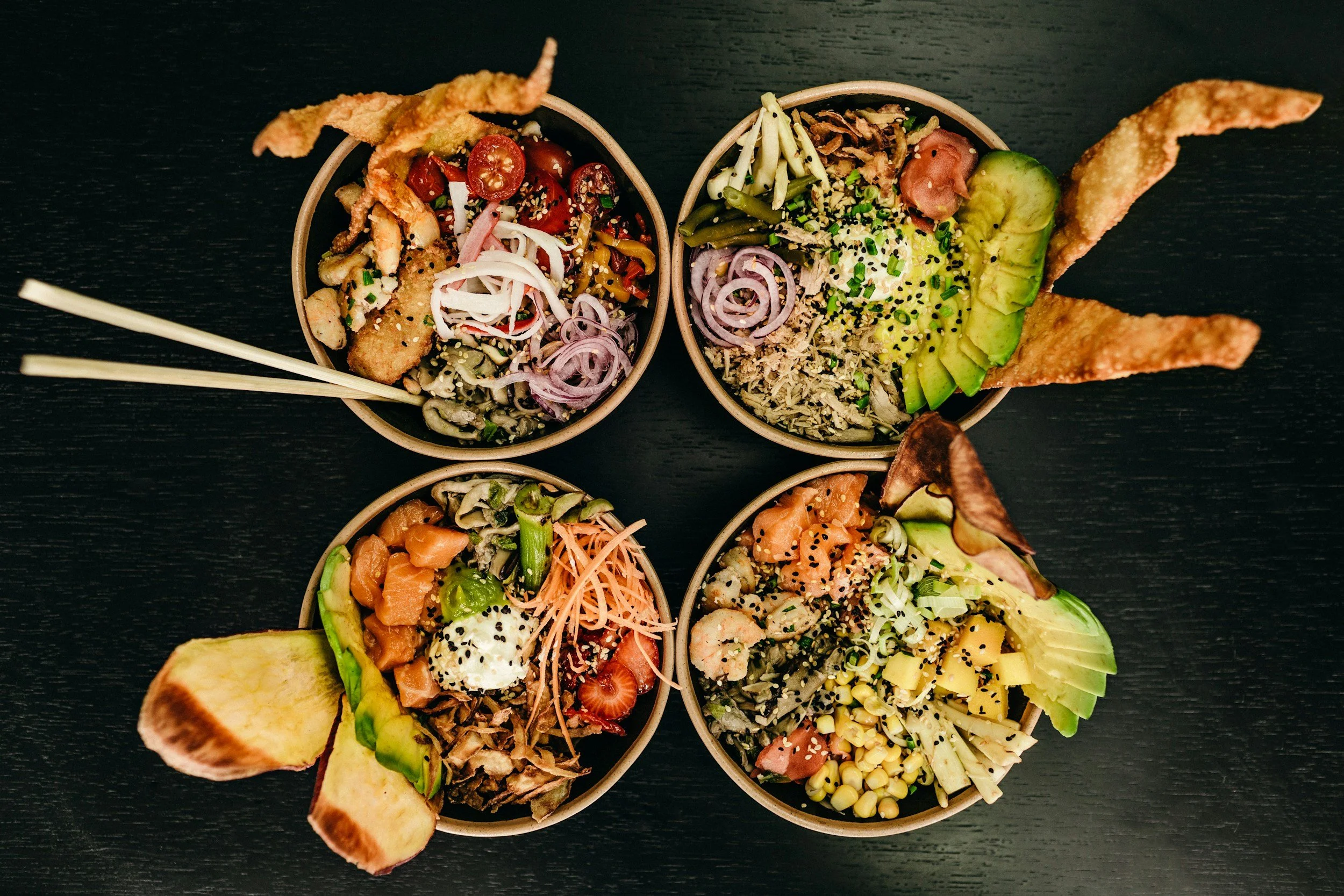 Four bowls of poke bowls with various sliced vegetables, seafood, and toppings, garnished with plantains and crispy chips on a black surface.