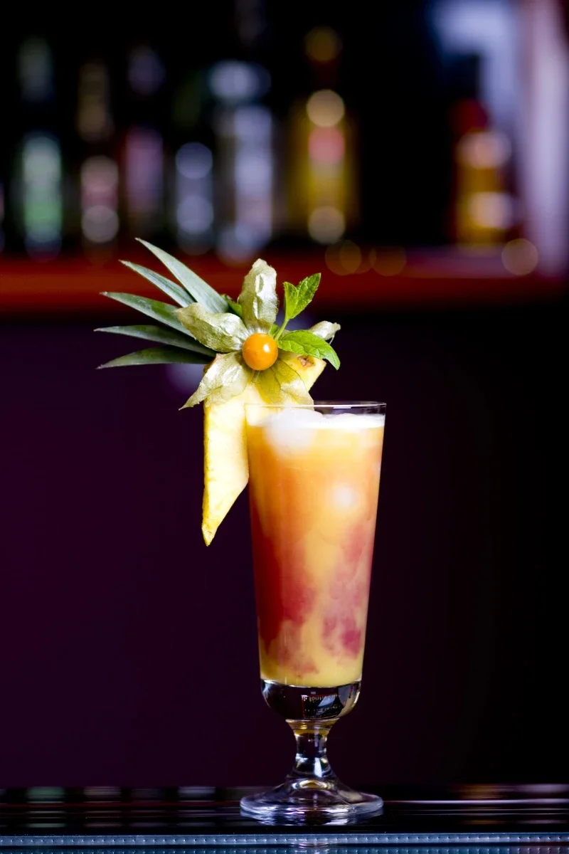 Tropical cocktail with pineapple, orange, and mint garnishes on a dark bar counter.