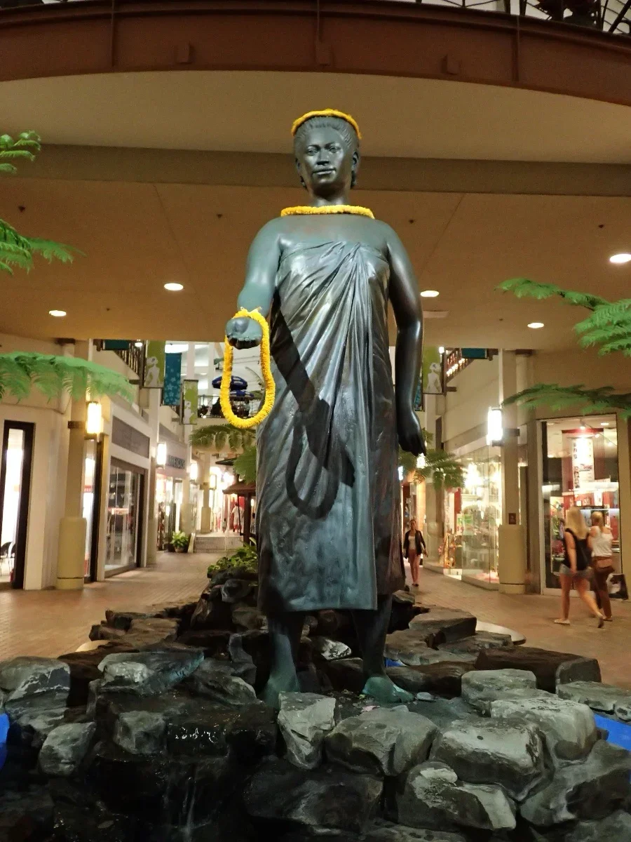 A bronze statue of a young girl dressed in traditional clothing, standing on rocks. The girl holds a ball in one hand, and is adorned with a yellow flower necklace and a flower headband. The statue is located inside a shopping mall or indoor shopping area, with storefronts and shoppers visible in the background.