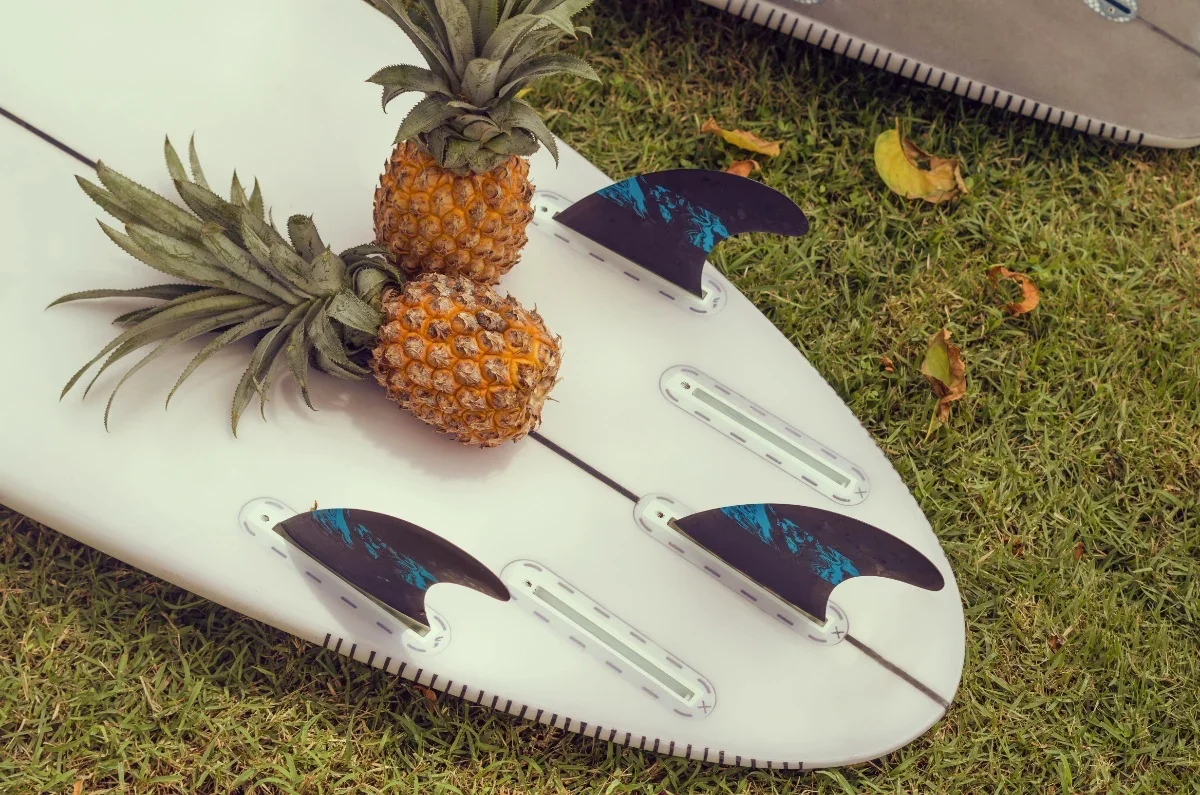 A surfboard lying on grass with two pineapples on top and surfboard fins attached.