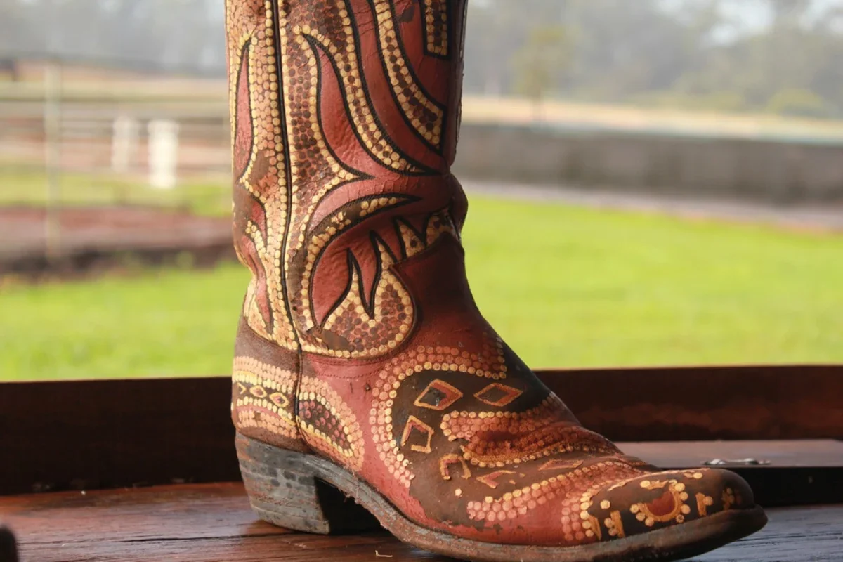 Close-up of a painted Western-style cowboy boot with intricate red, black, and yellow designs, placed indoors on a wooden surface, with a blurred outdoor background.