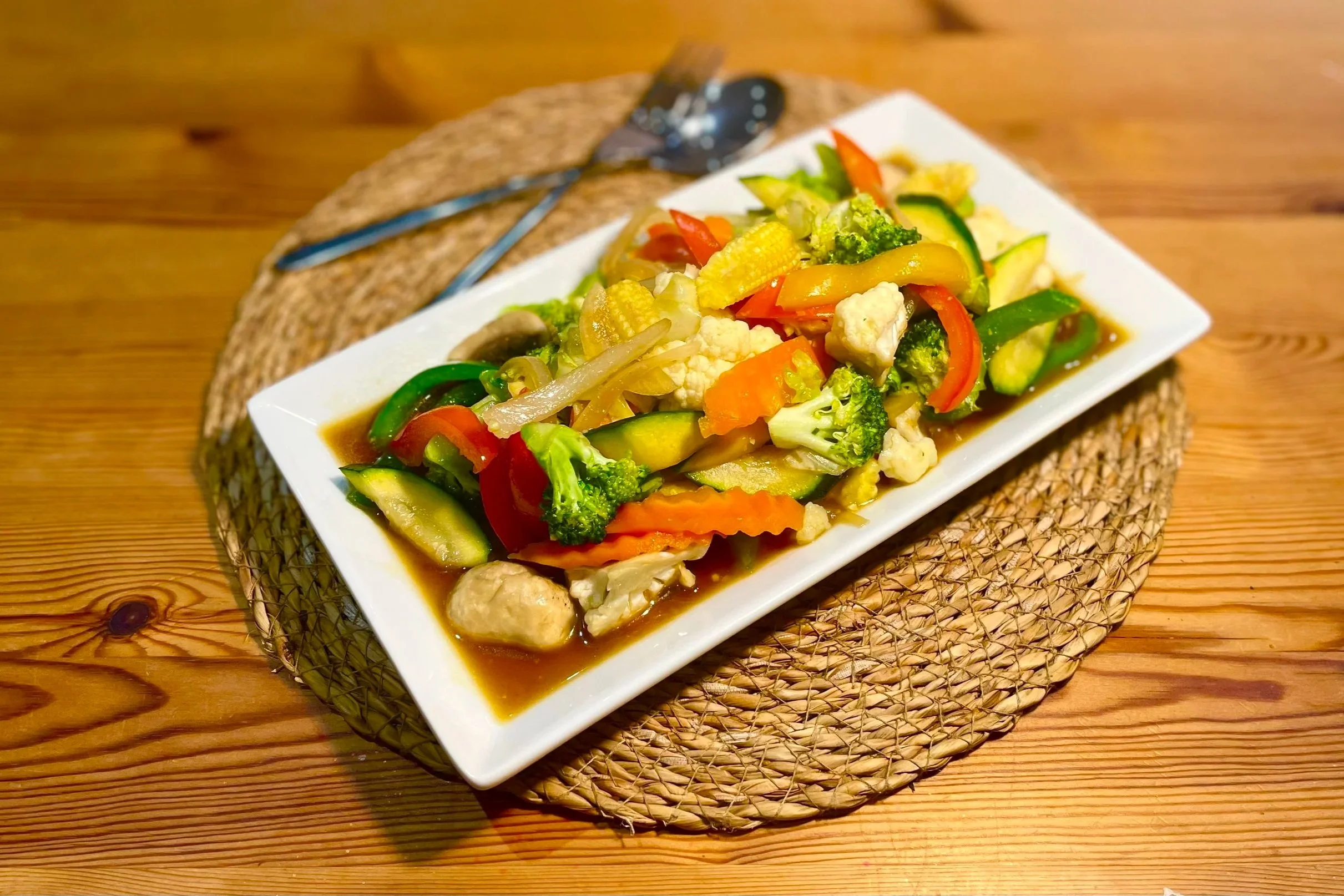 A plate of stir-fried mixed vegetables with broccoli, carrots, bell peppers, zucchini, cauliflower, and onions in sauce, set on a woven placemat on a wooden table.