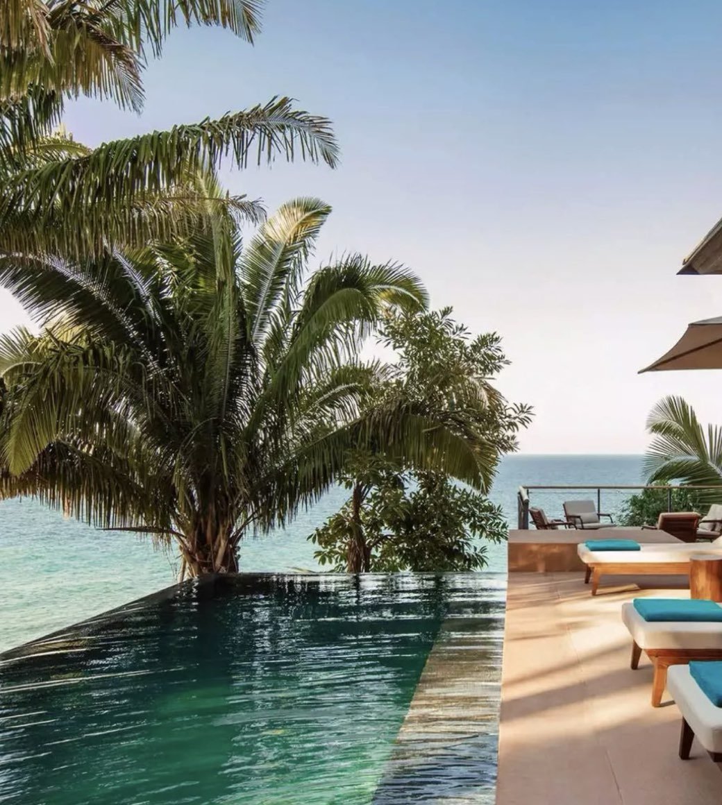 A coastal scene featuring an infinity pool overlooking the ocean, with palm trees, outdoor seating, and umbrellas on a deck.