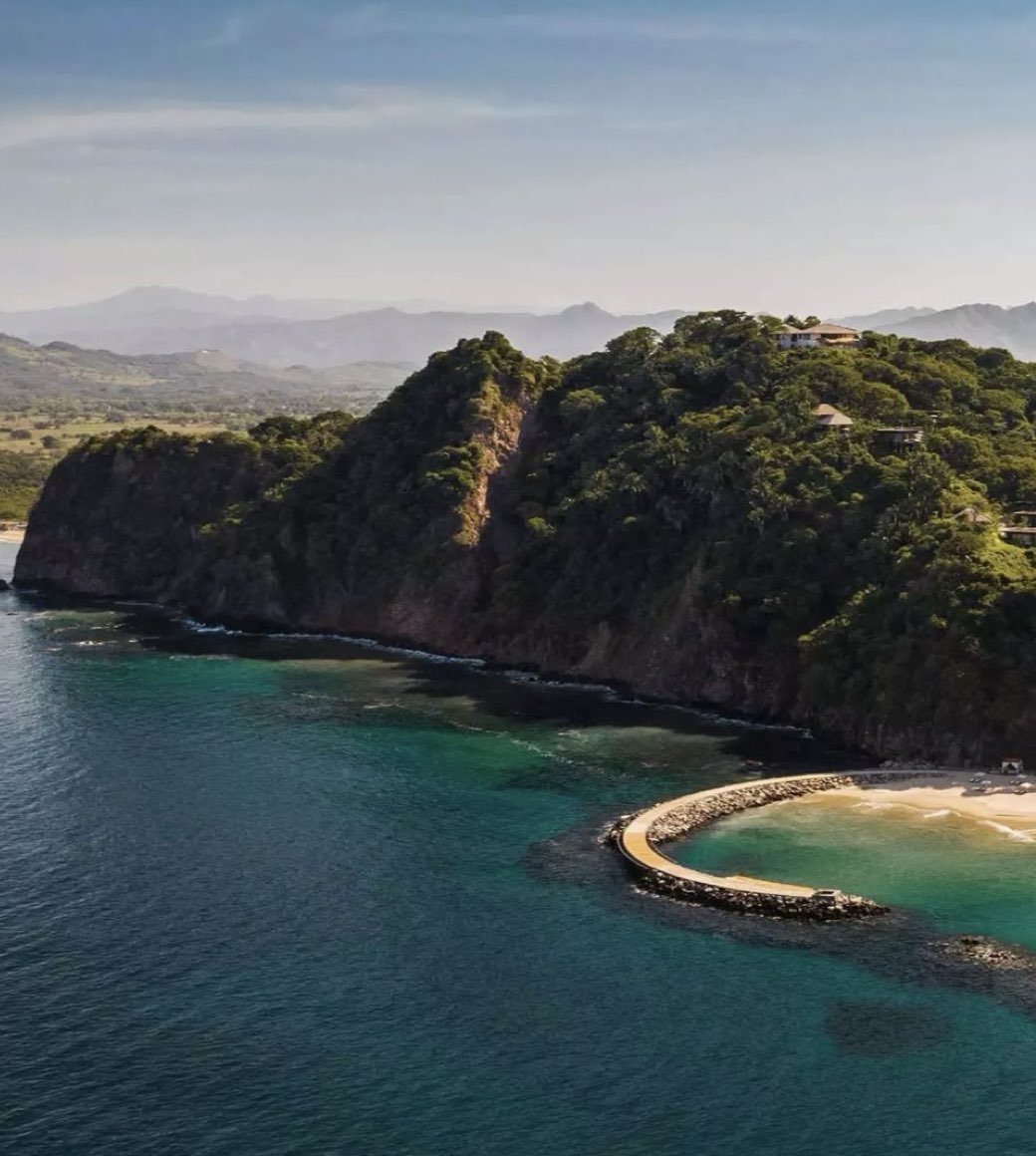 Coastal landscape with green hills, a curved breakwater, and a sandy beach, overlooking the ocean and distant mountains.