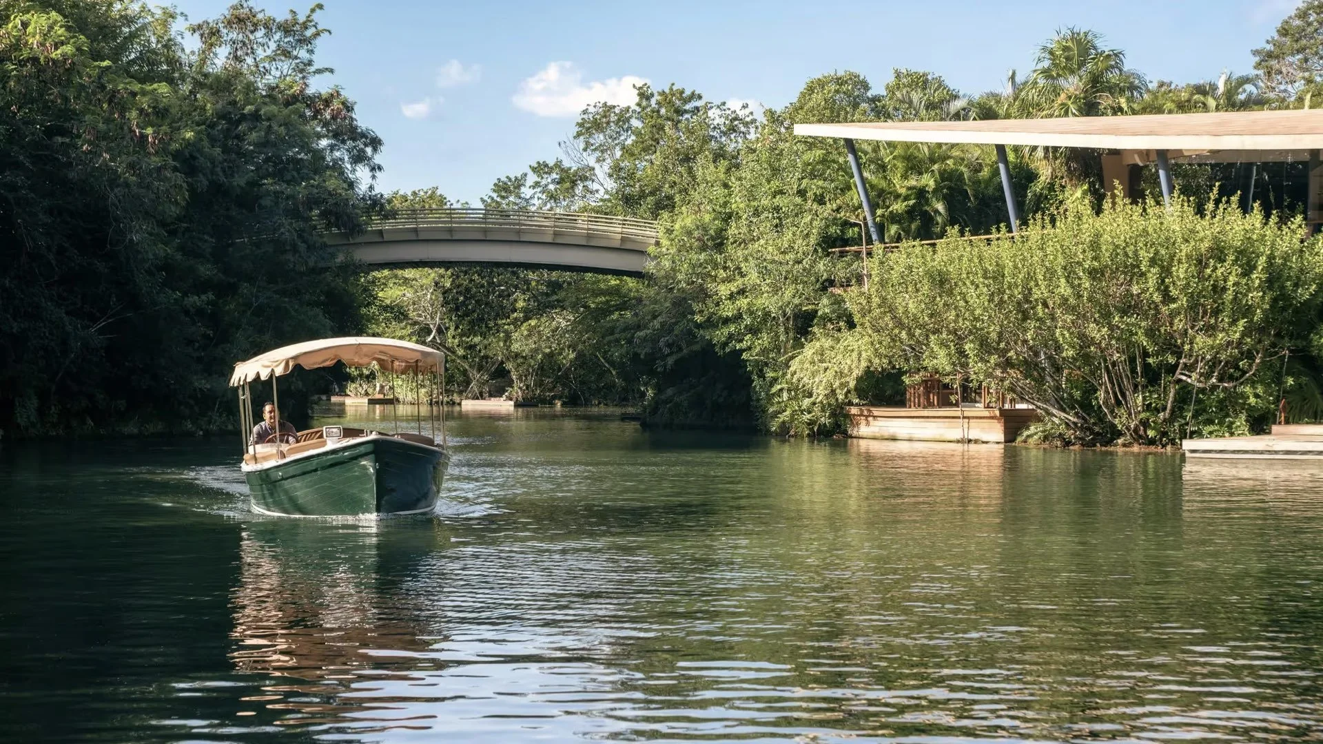 A boat with a beige canopy on a calm river, surrounded by lush green trees and bushes, with a modern bridge and building in the background under a blue sky with a few clouds.