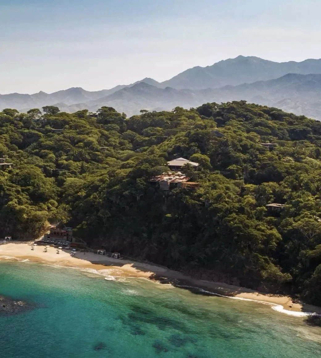 Aerial view of a lush green hillside near a beach with cabins and buildings, overlooking turquoise ocean waters with mountains in the background.