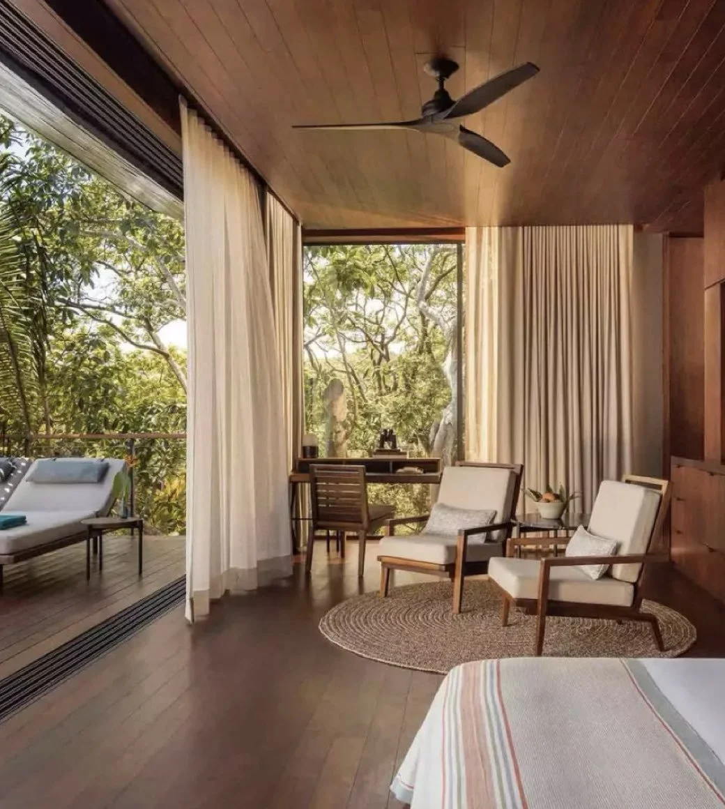 A cozy living room with large windows showing lush greenery outside, wooden ceiling, beige curtains, two white armchairs with wooden frames and pillows, a small round woven rug, a wooden desk with a chair, a ceiling fan, and a sliding glass door lead