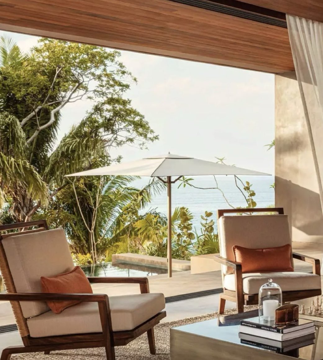 Living room with two armchairs, a coffee table with books and a glass dome, and an outdoor view of trees, a white umbrella, and the ocean.