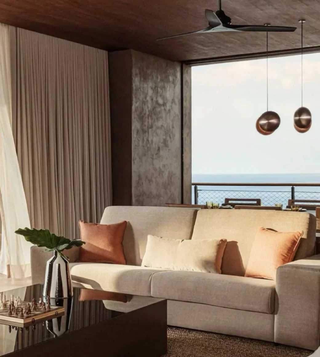 Living room with beige sofa, three cushions in shades of orange and beige, a metal vase with a large green leaf on a coffee table, a chessboard, a window with a view of the ocean, beige curtains, wooden ceiling, textured wall, modern ceiling fan, and